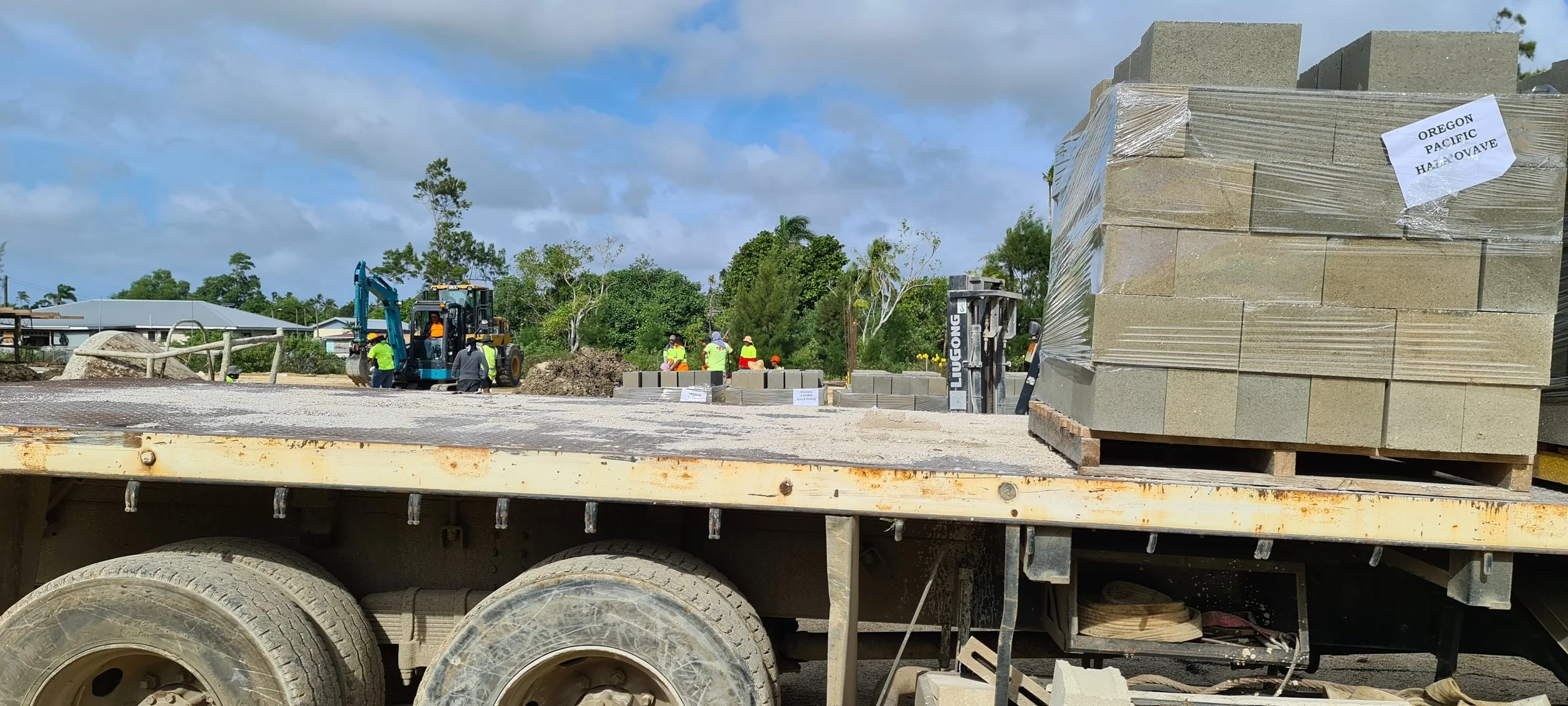 Construction workers in safety vests and helmets working on a building site, with a large stack of concrete blocks on a truck and construction machinery in the background.