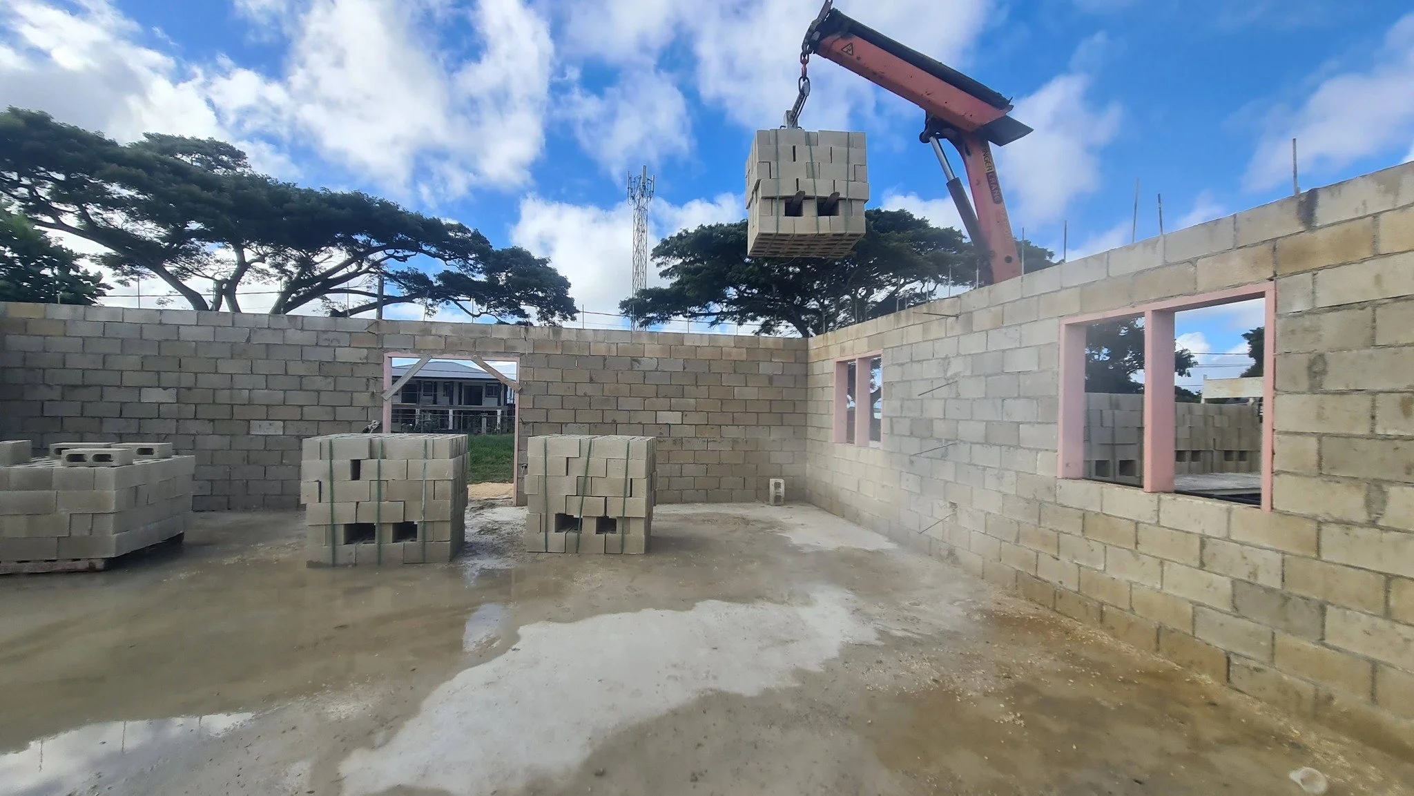 Construction site with brick walls and windows being built, a crane lifting a pallet of bricks, and a cloudy sky with trees in the background.
