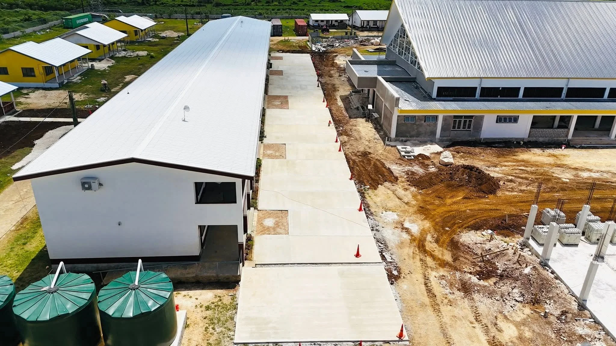 Aerial view of a construction site with buildings, some with metal roofing and painted in yellow, and a paved walkway with orange cones. There are also three green water tanks and dirt areas with construction materials.