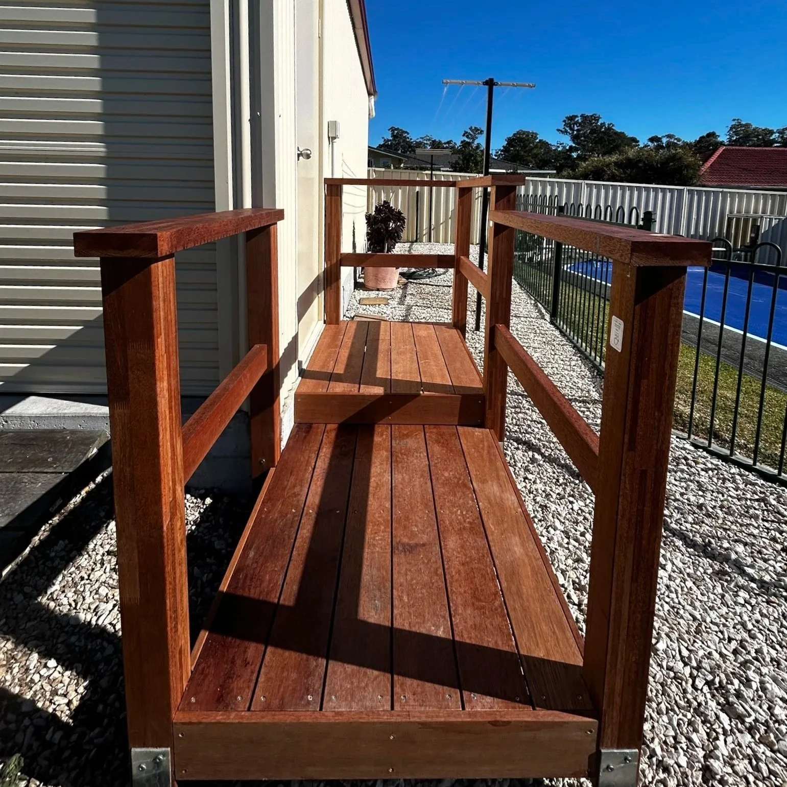 A wooden ramp with railings on both sides, leading up to a door outside of a shed, surrounded by gravel and fencing, under a clear blue sky.