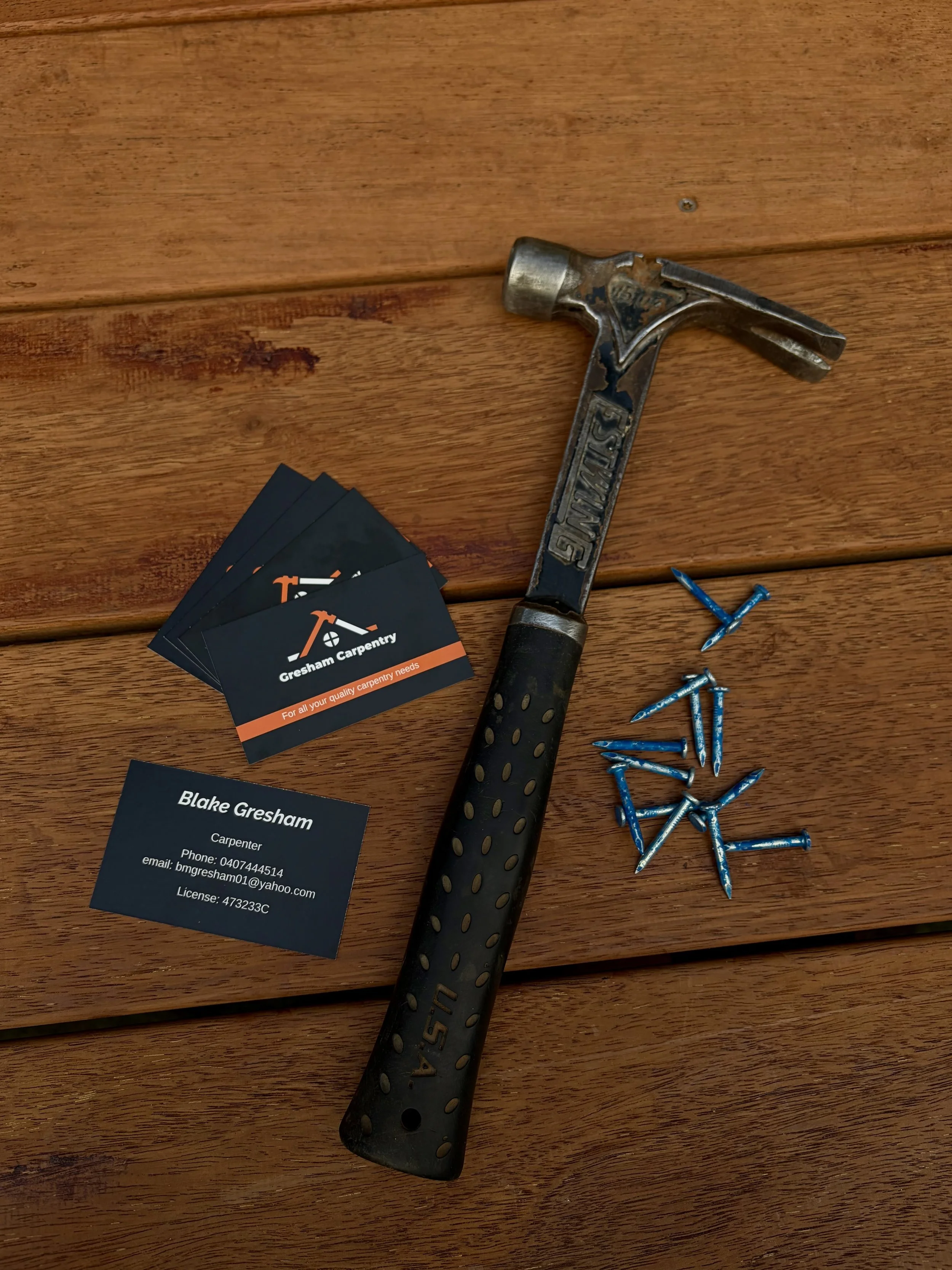 A hammer with a black rubber grip and the word "STANLEY" on the side, a business card with contact information for Blake Gresham, a pile of blue screws, and some black and orange cards for Gresham Carpentry on a wooden surface.