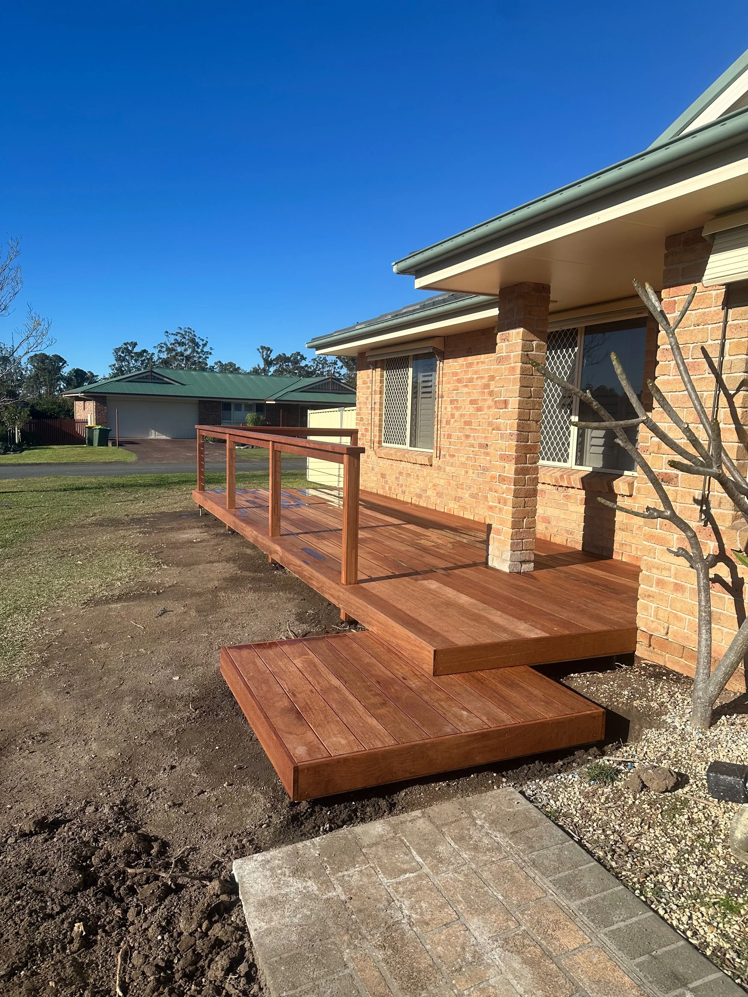 A newly built wooden deck attached to a brick house under a clear blue sky. The deck has a railing and extends from the house to the yard. There is a custom step to create easy access to the deck.