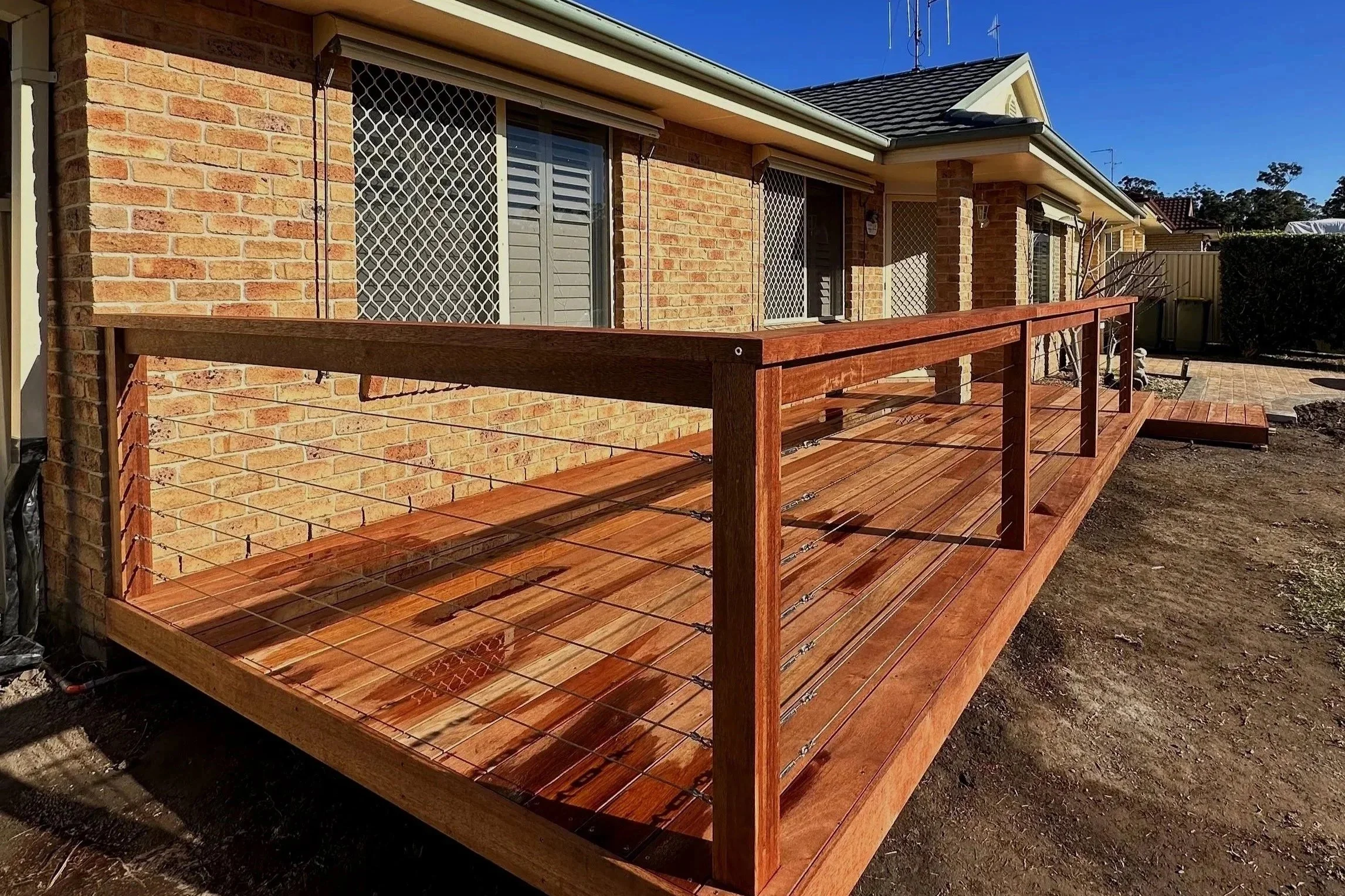 Wooden deck attached to brick house with large windows, white security bars, and a privacy shutter, under a gray roof with antennas, in a sunny yard with some plants and a brick paver area.
