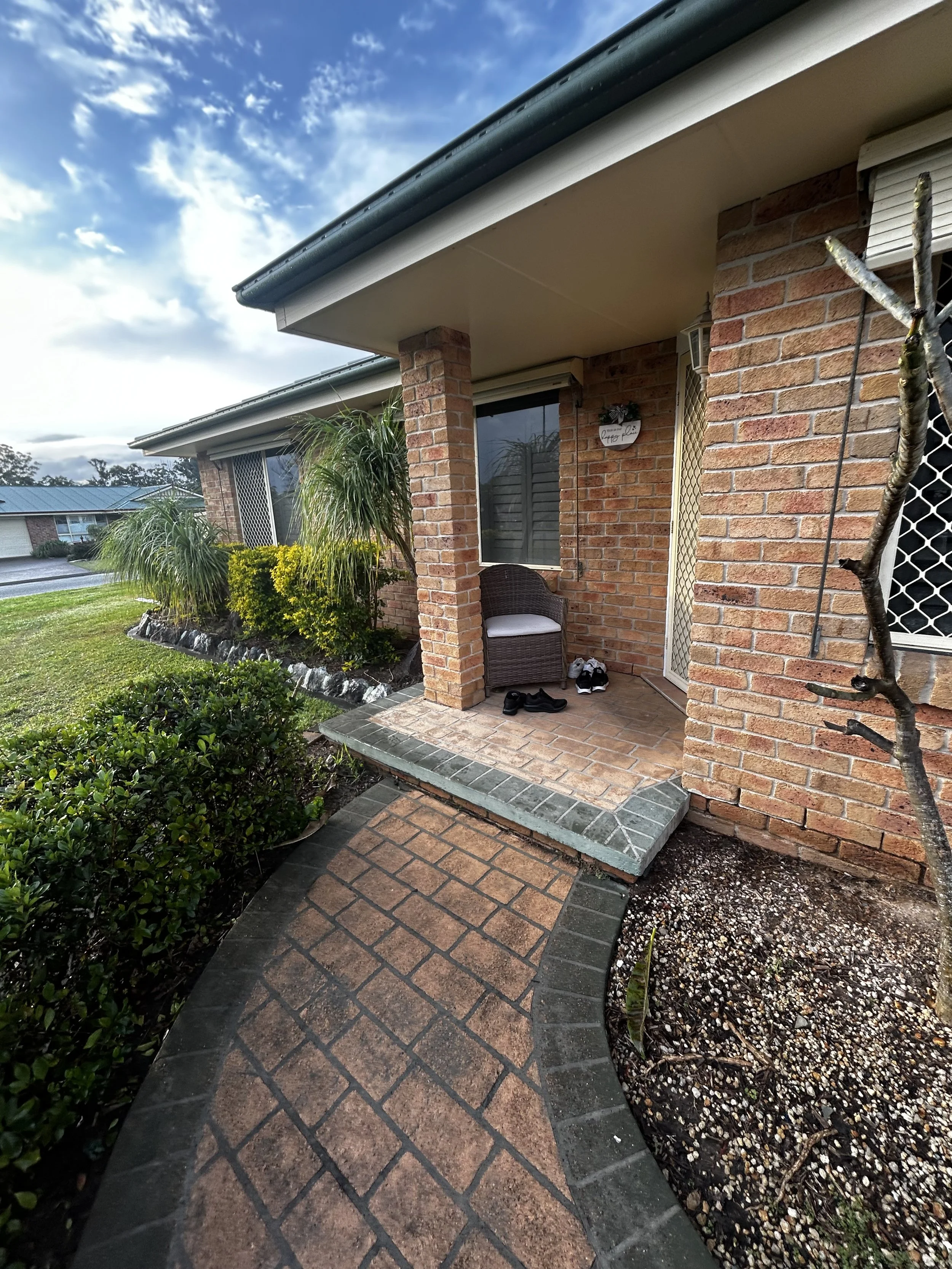 The image shows the front porch of a brick house with a tiled walkway leading up to it. There are plants and bushes in the garden area surrounding the walkway. This is the before project.