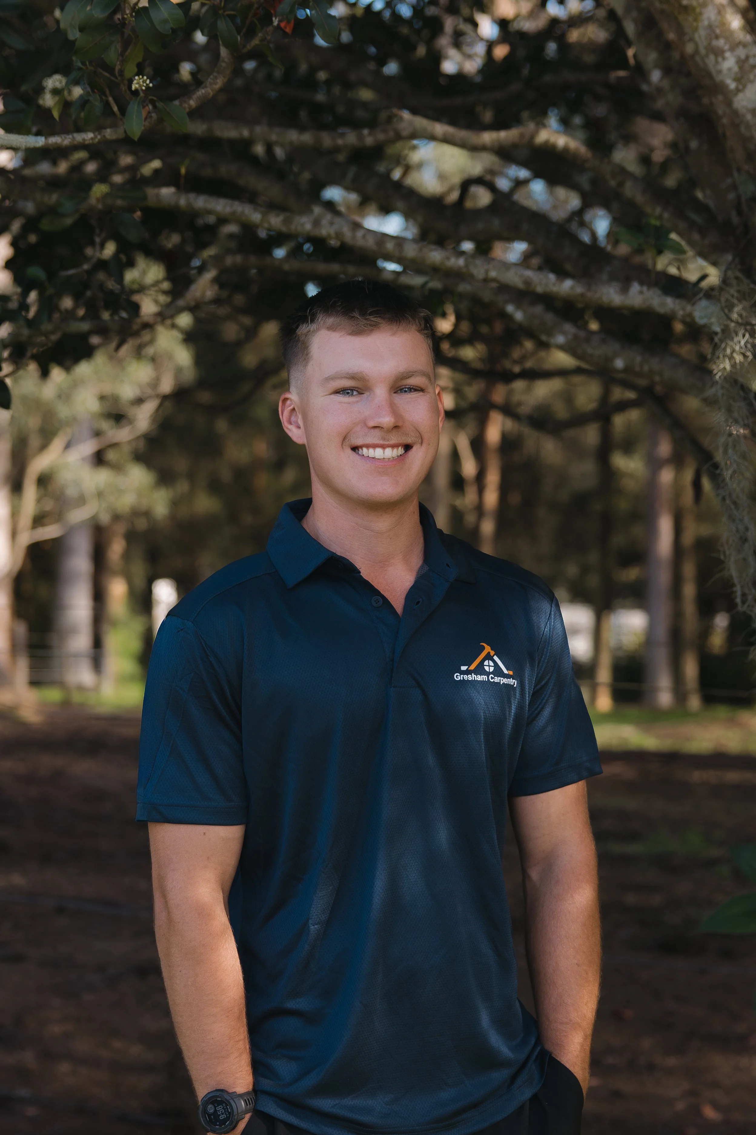 Young business owner, Blake Gresham wearing a navy blue polo shirt with a company logo that reads 'Gresham Carpentry' and a wristwatch.
