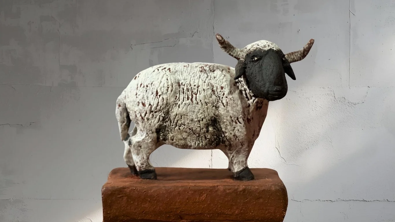 Handgefertigte Skulptur eines Bullen auf einem braunen Podest, mit schwarzem Gesicht und weißen krakelierten Körper, vor einem grauen Hintergrund.