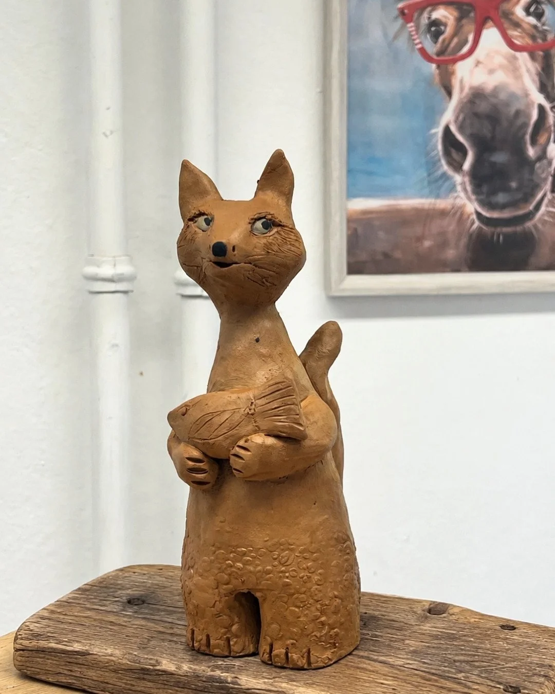 Eine Keramikfigur eines Katzenkopfes, die eine Muschel hält, auf einem Holzsockel. Im Hintergrund ist ein Poster eines Hundes, der Sonnenbrille trägt.
