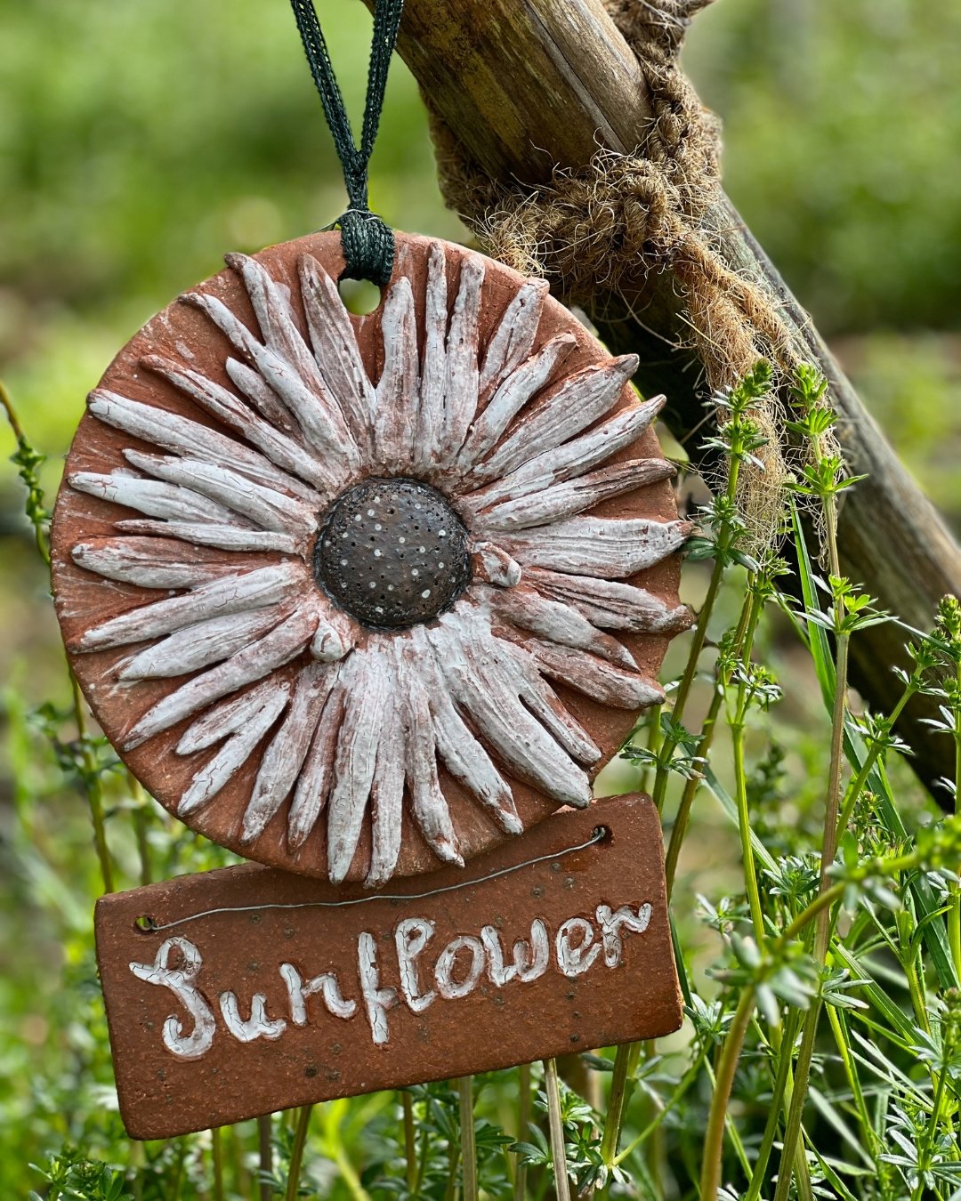 Ein Keramikschild in Form einer Sonnenblume mit einer kleinen Tafel darunter, auf der 'Sunflower' steht, hängt an einem Ast im Garten, umgeben von grünen Pflanzen.