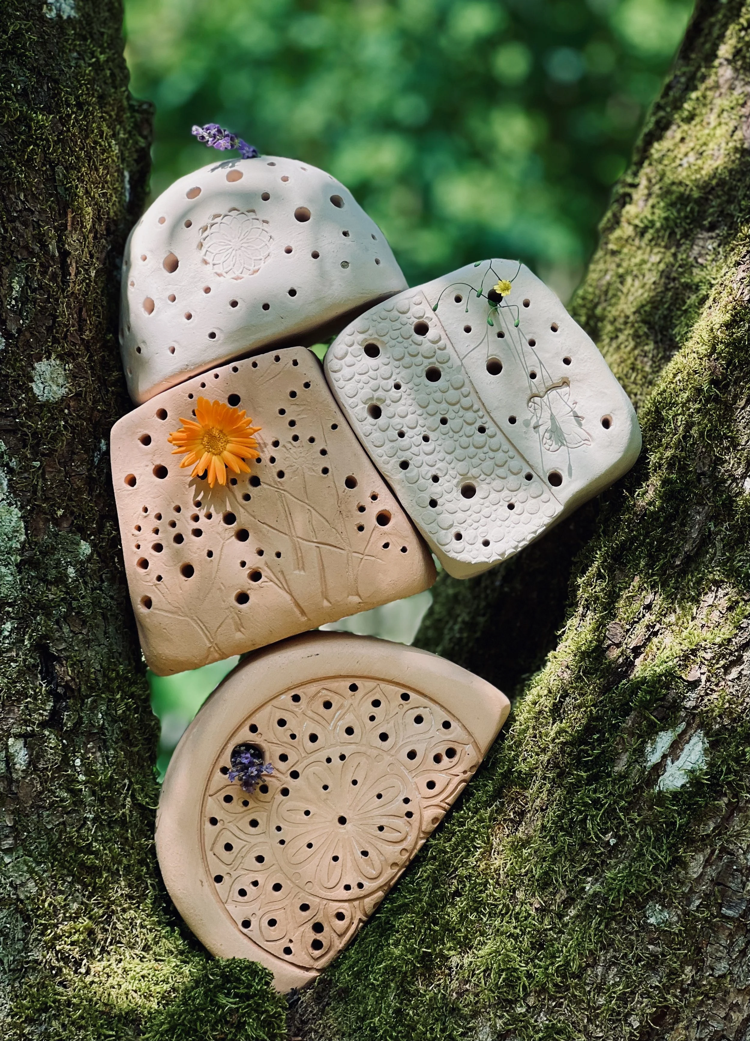 Mehrere handgefertigte Keramikplatten mit floralen und geometrischen Mustern, dekorativ an einem Baumstamm mit Moos angebracht, einige mit Blumen und Lavendel verziert.