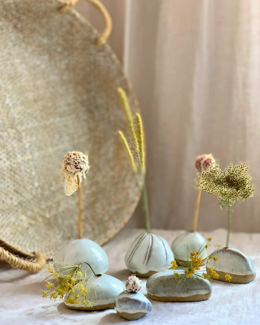Deko mit weißen Steinen, Trockenblumen und keramischen Vasen vor einem großen, runden, goldfarbenen Gegenstand und einem weißen Vorhang im Hintergrund.