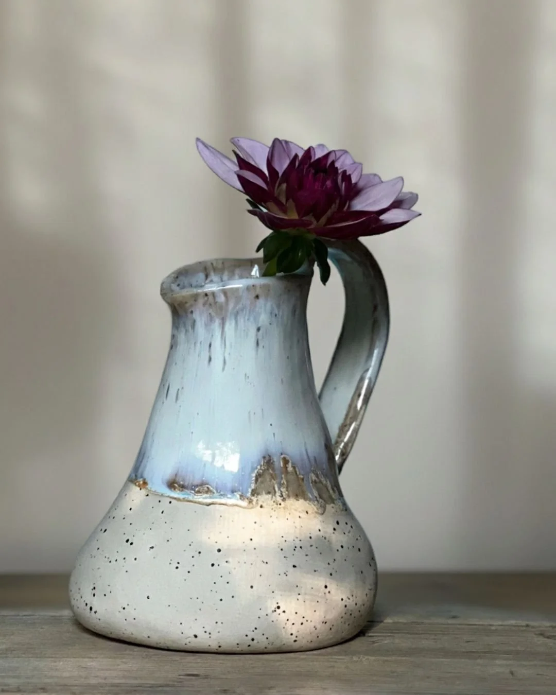 Auf diesem Bild ist eine Keramikvase mit einem Stiel, die einen bunten Blumenstrauß mit einer großen, rosa-lila Blume enthält, vor einem neutralen Hintergrund.