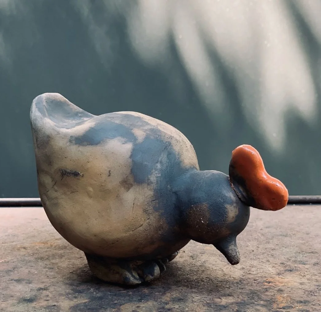 Ein bemalter Tonvogel mit großem orangefarbenem Schnabel auf einem Holztisch, im Hintergrund unscharf.