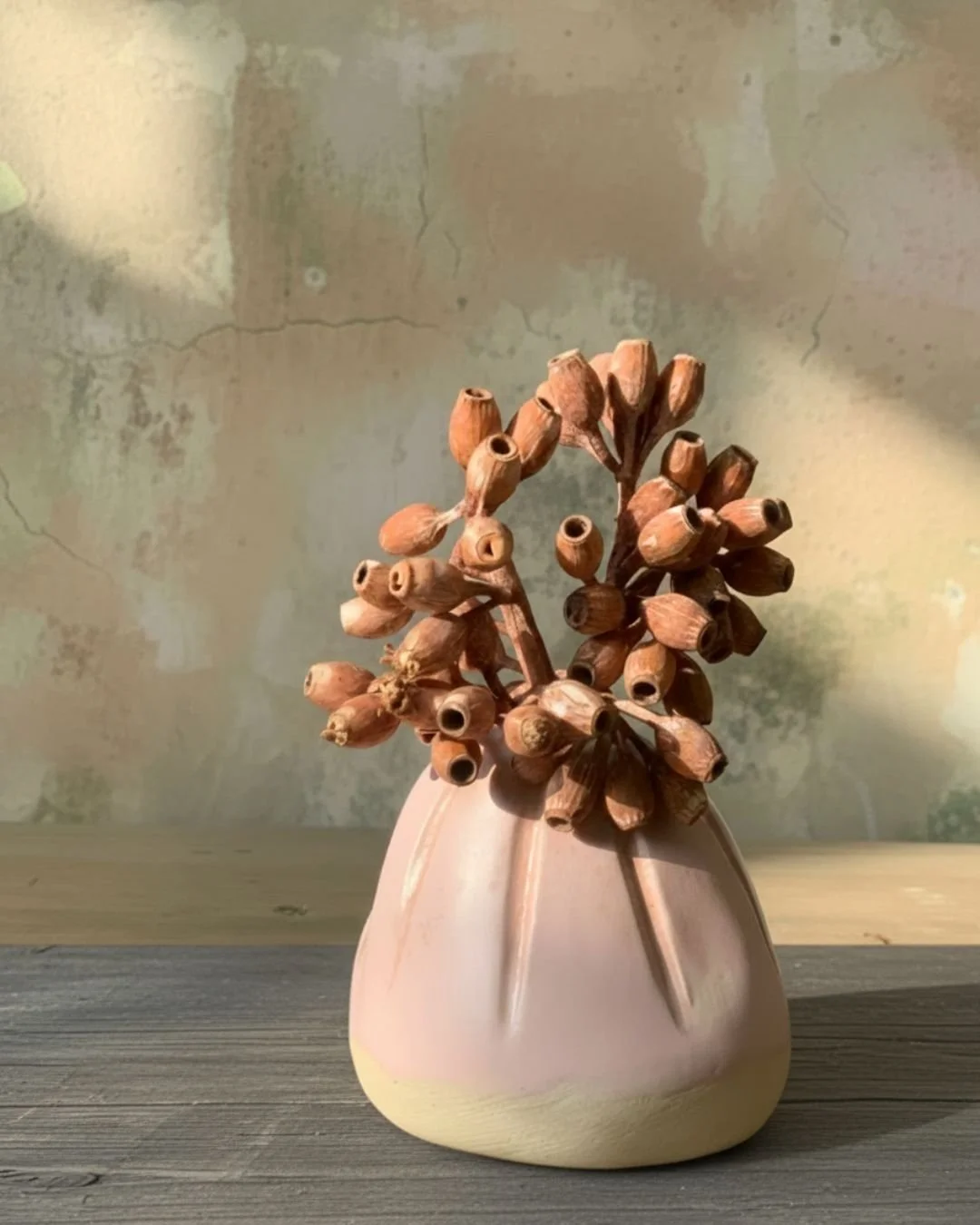 Kunstvolle Vase mit getrockneten Pflanzen auf Holztisch vor einer gemalten Wand.