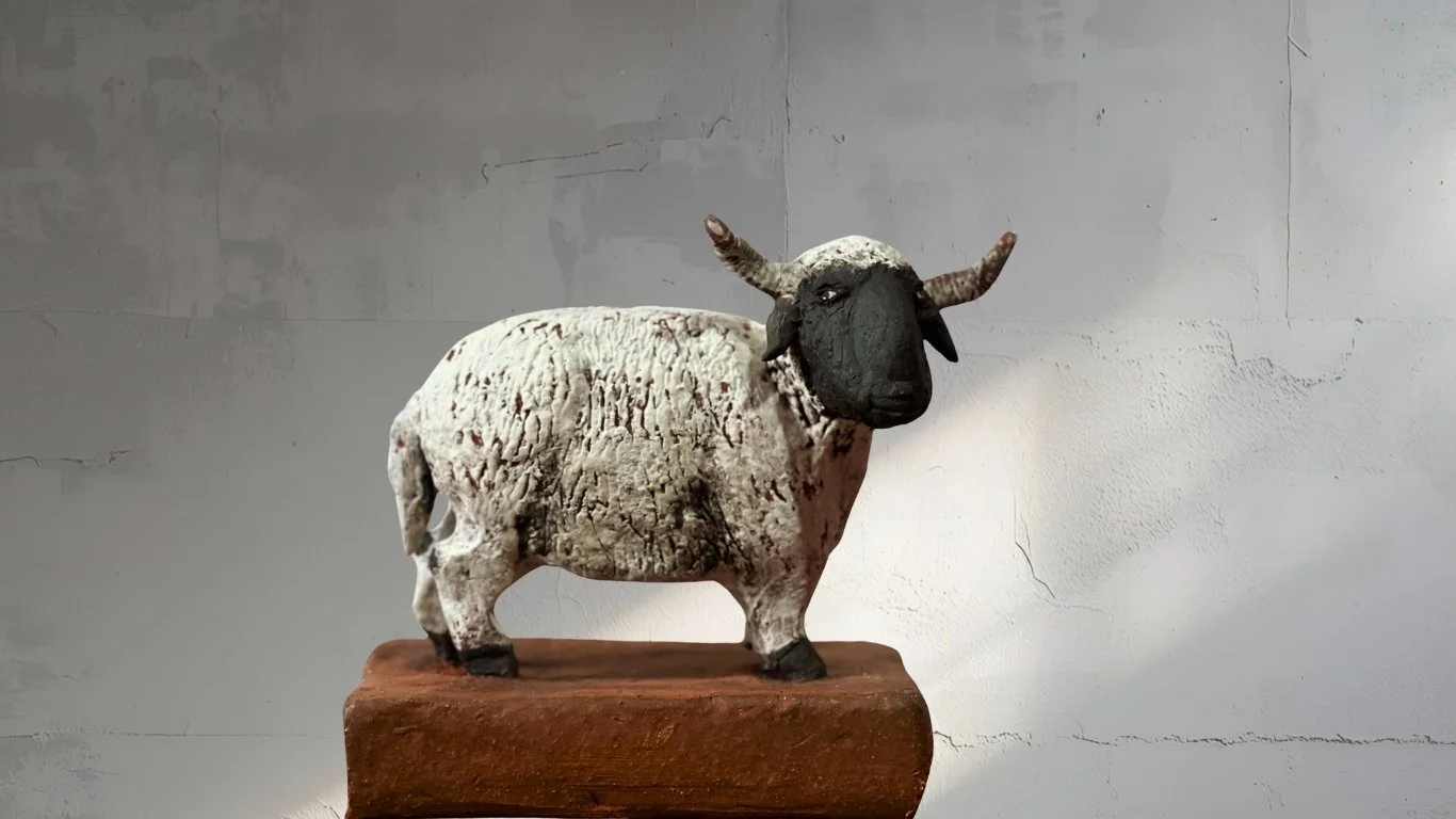 Kunststofffigur eines Schafes mit schwarzem Kopf und Hörnern, steht auf einem Holzsockel vor einer weißen Wand.