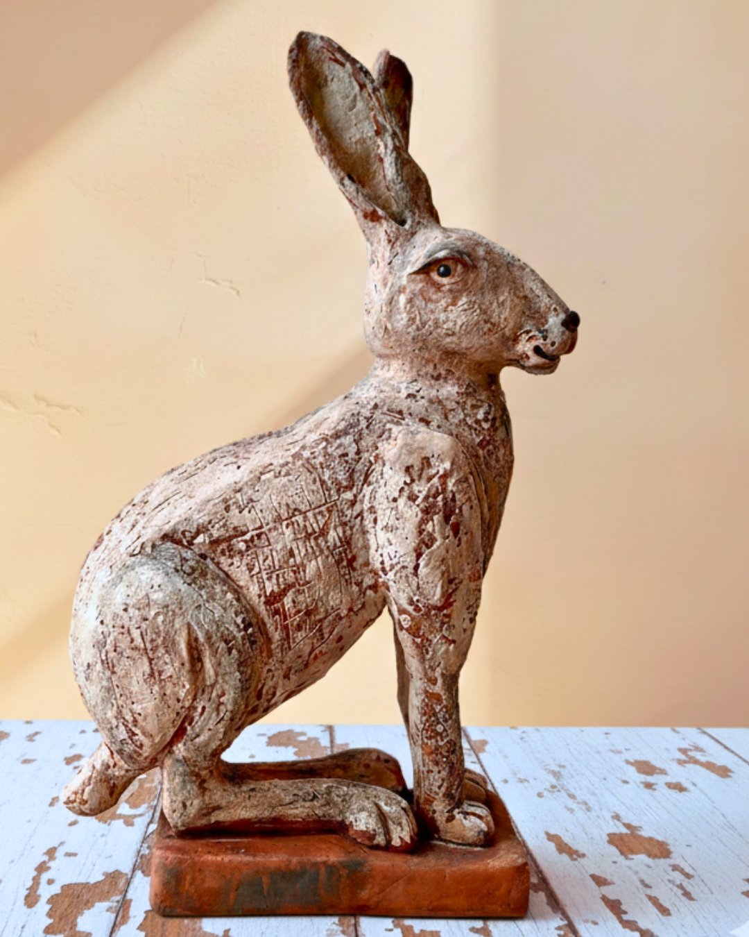 Alter bemalter Holzschnitzfigur eines Hasen mit langen Ohren, sitzend auf einem Holzsockel auf einem alten, abgenutzten Tisch.