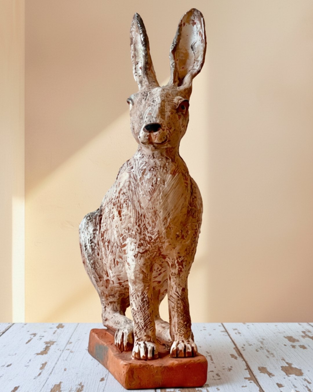 Holzfigur eines sitzenden Hasen mit großen Ohren auf einem weißen, abgenutzten Holztisch, vor einem beige-hellen Hintergrund.