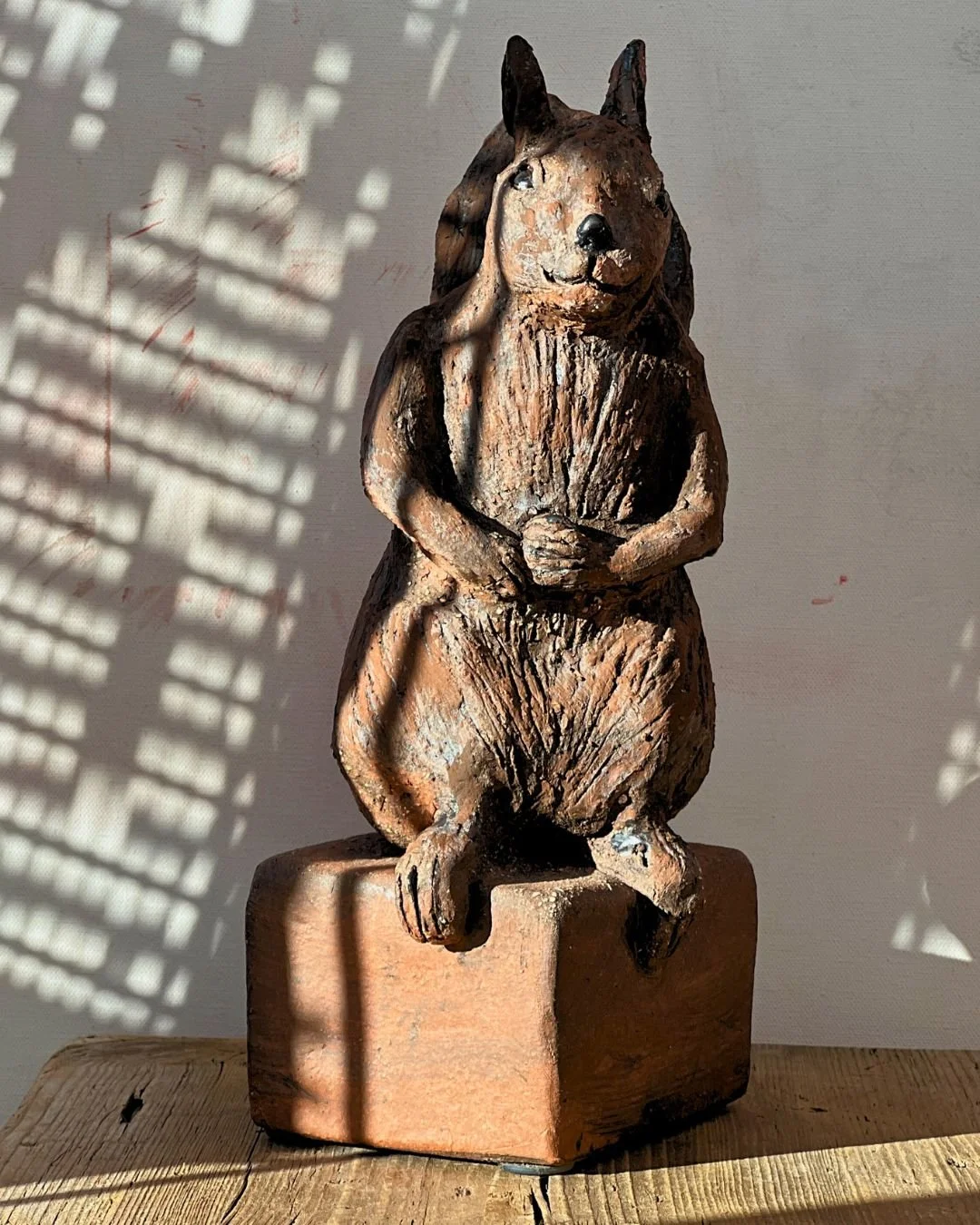 Bronzefigur eines sitzenden Eichhörnchens mit Händen im Schoß, auf einem rechteckigen Sockel, mit Schatten an der Wand im Hintergrund.