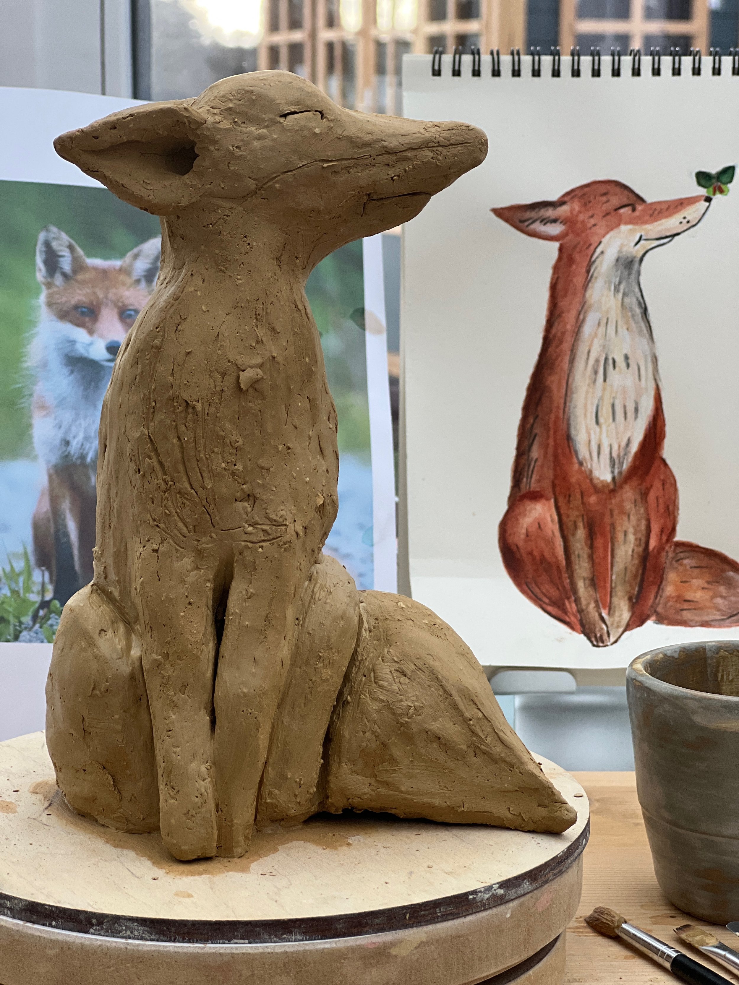 Eine Terrakotta-Figur eines sitzenden Hundes mit geschlossenen Augen, auf einem Drehteller in einem Kunststudio. Im Hintergrund sind Fotos von Tieren sowie eine Skizze eines Fuchses mit einer Blattattrappe zu sehen.
