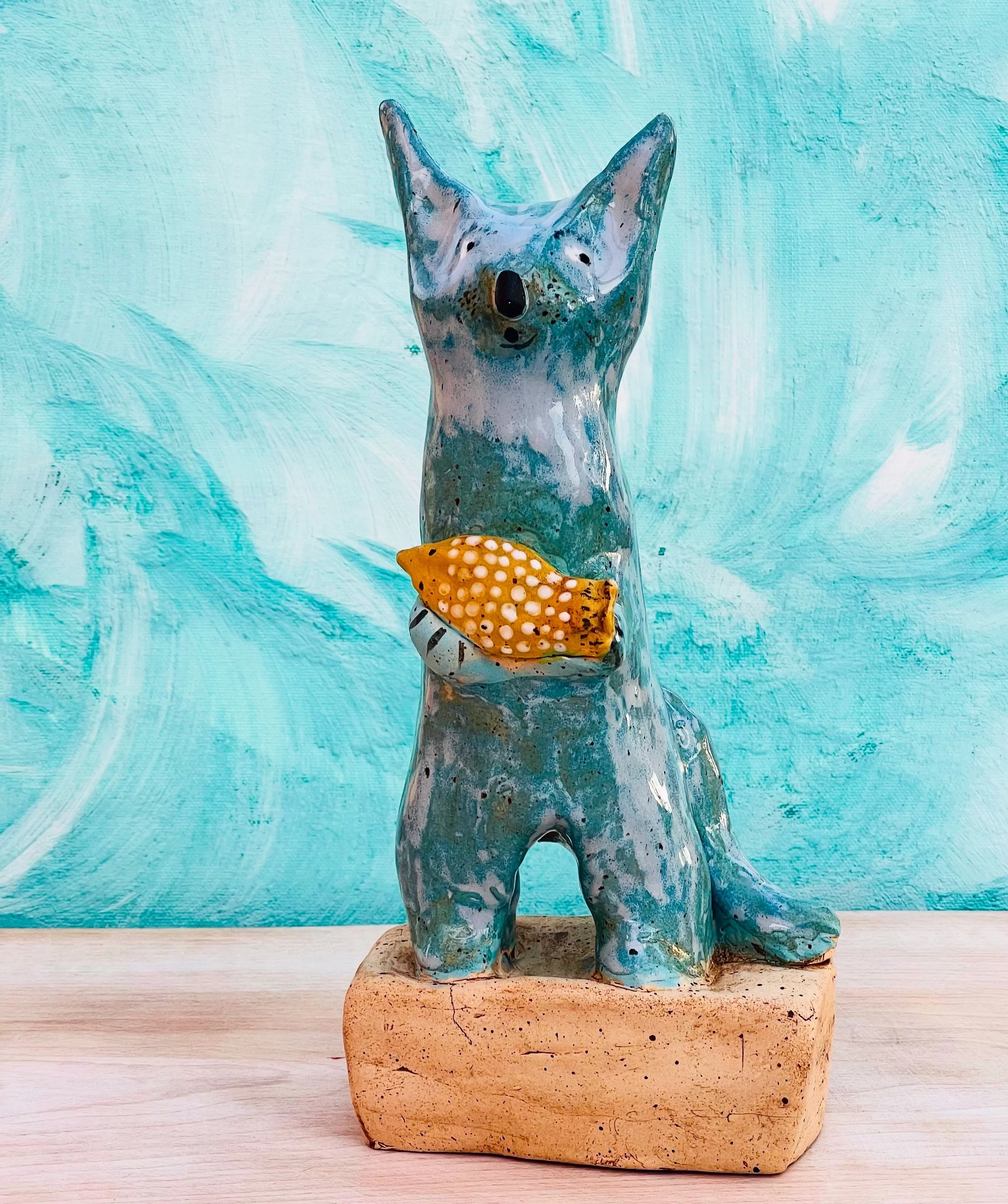 Kunststofffigur eines Hundes mit großen Ohren, die auf einem rechteckigen Sockel steht, auf einem hellen Holztisch vor einem aquafarbenen Hintergrund.