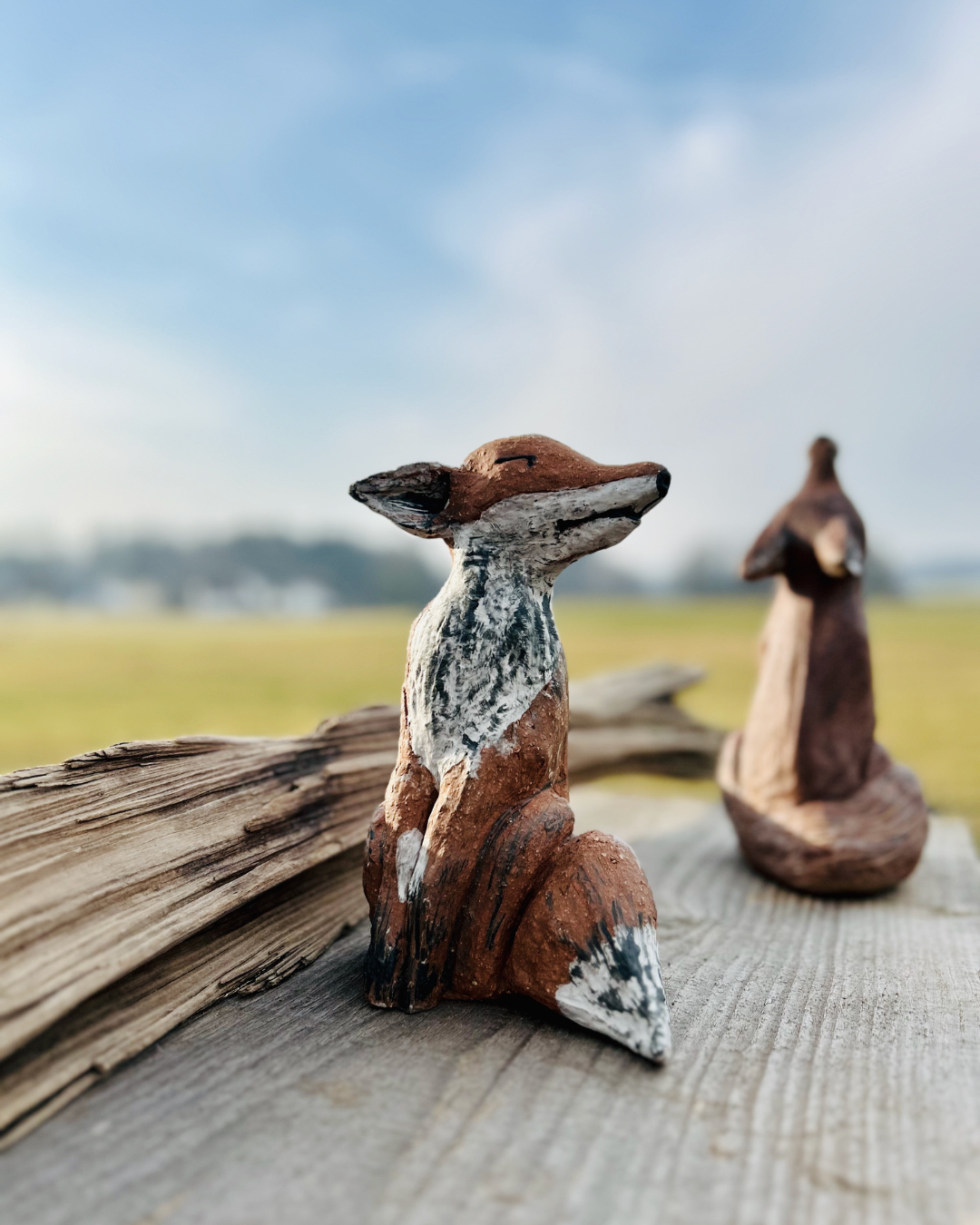 Zwei handgefertigte Tierfiguren aus Holz auf einem Holzbrett, mit einer ländlichen Landschaft im Hintergrund.