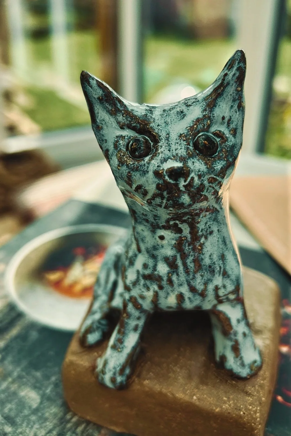 Porzellanskulptur eines sitzenden Hundes auf Holzbasis, mit patiniertem bläulichen Finish, im Hintergrund ein Fenster und eine Tasse auf einem Tischtuch.