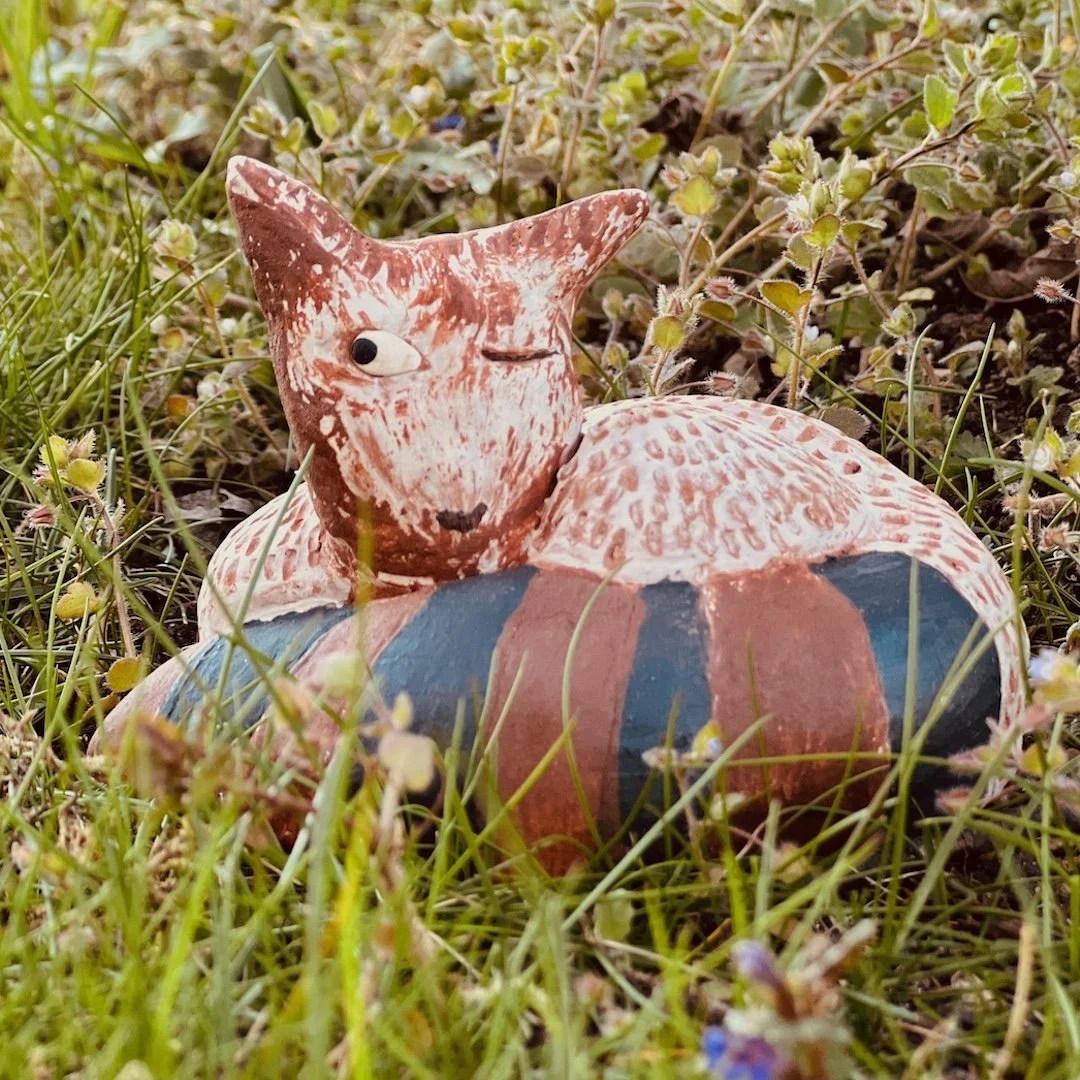 Eine bemalte, terrakottaartige Katze-Figur liegt im Gras mit kleinen Blumen.