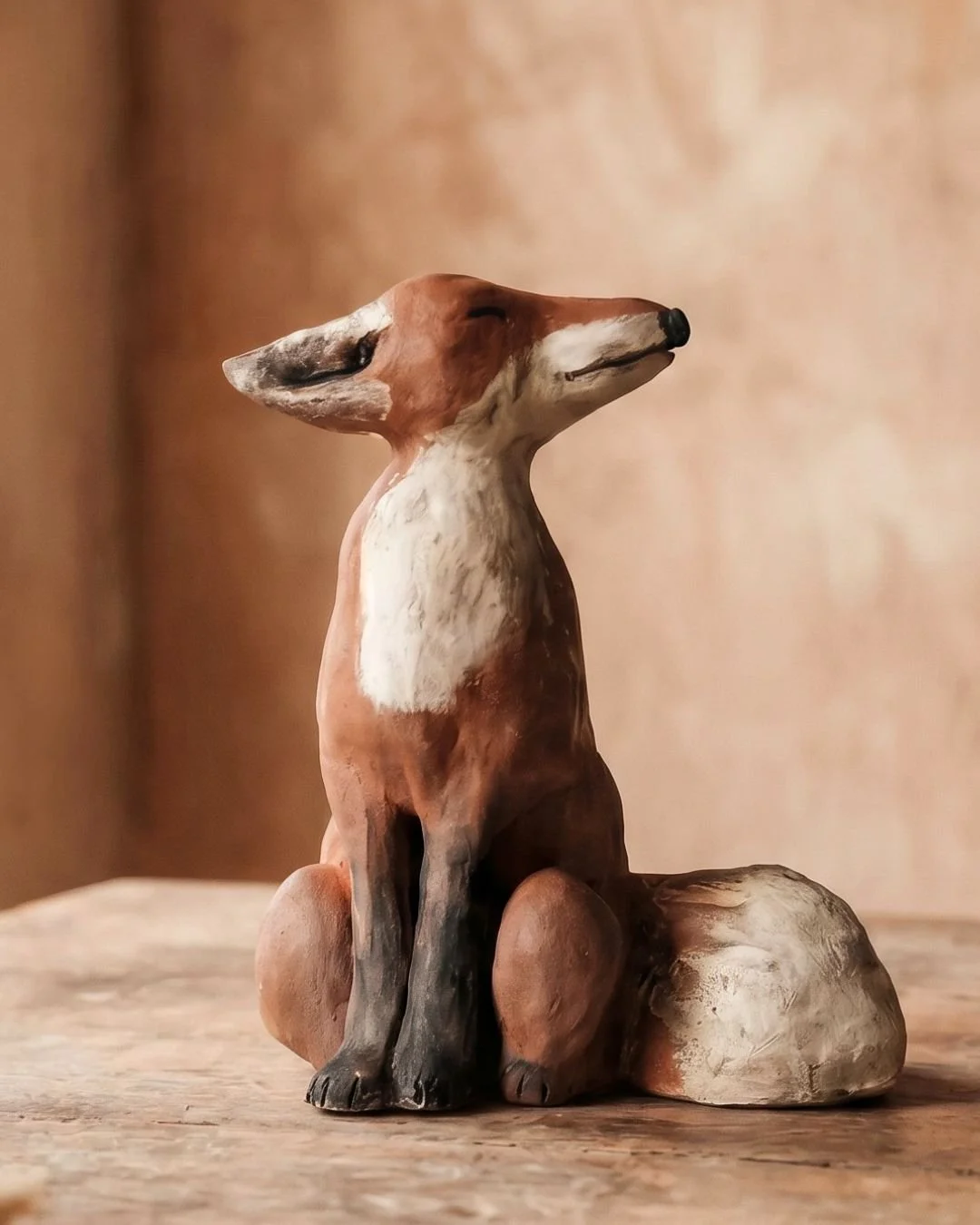 Ein handgefertigtes, bemaltes Keramik- oder Tonfigur eines sitzenden Fuchses auf Holzoberfläche vor einem braunen, verwaschenen Hintergrund.