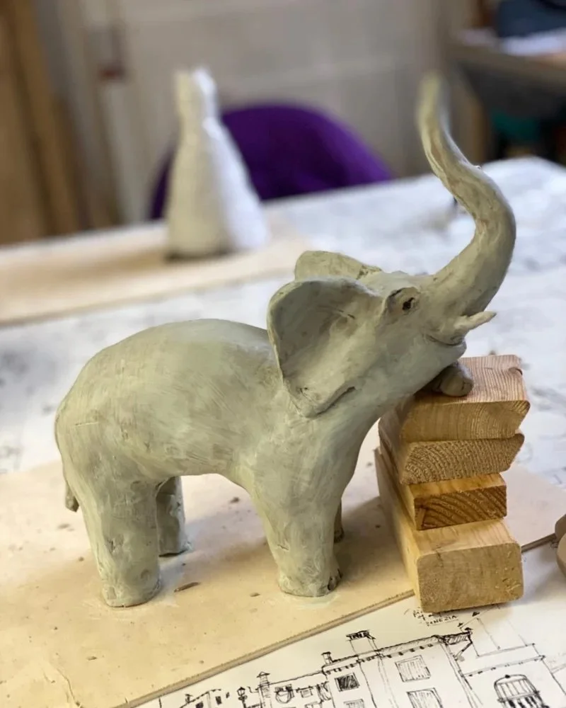 Eine handgefertigte Elefantenfigur aus Holz auf einem Tisch, neben einem kleinen Holzstapel und einem Architekturplan.
