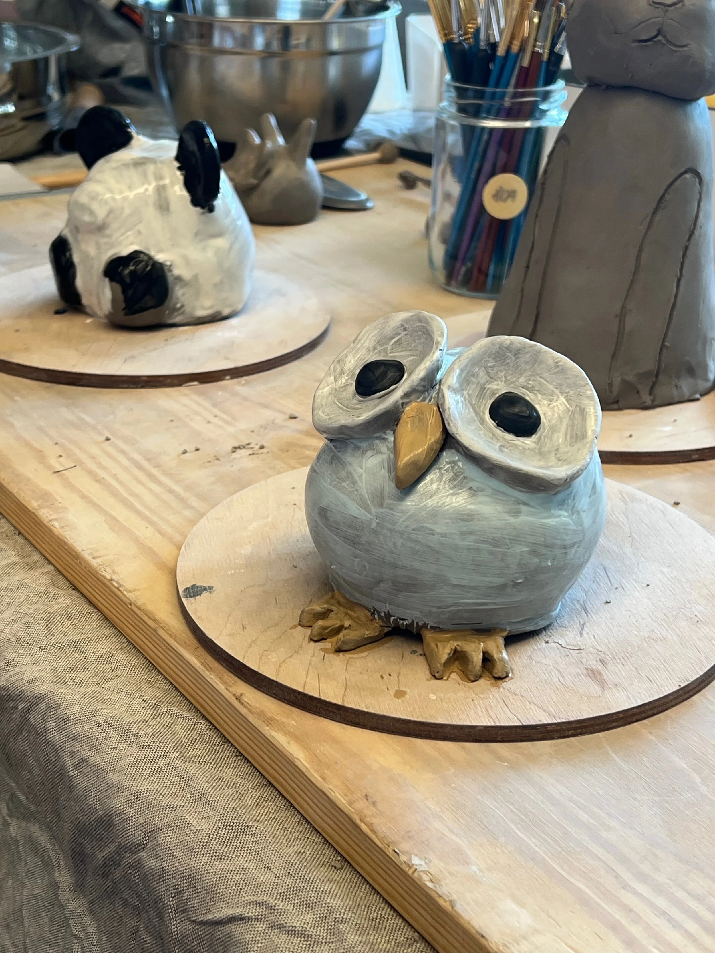 Keramikskulpturen in Form von Tieren auf einem Holztisch, darunter eine Eule, ein Schwein und Hase, mit Pinsel in einem Glasbehälter im Hintergrund.