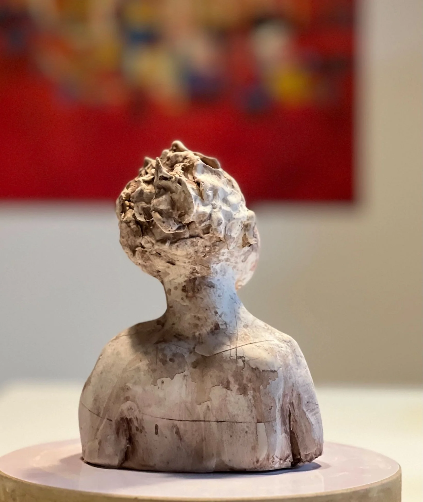 Skulptur eines menschlichen Kopfes und Oberkörpers aus gebrochenem, hellen Stein auf einer runden Plattform, im Hintergrund eine bunte Wand