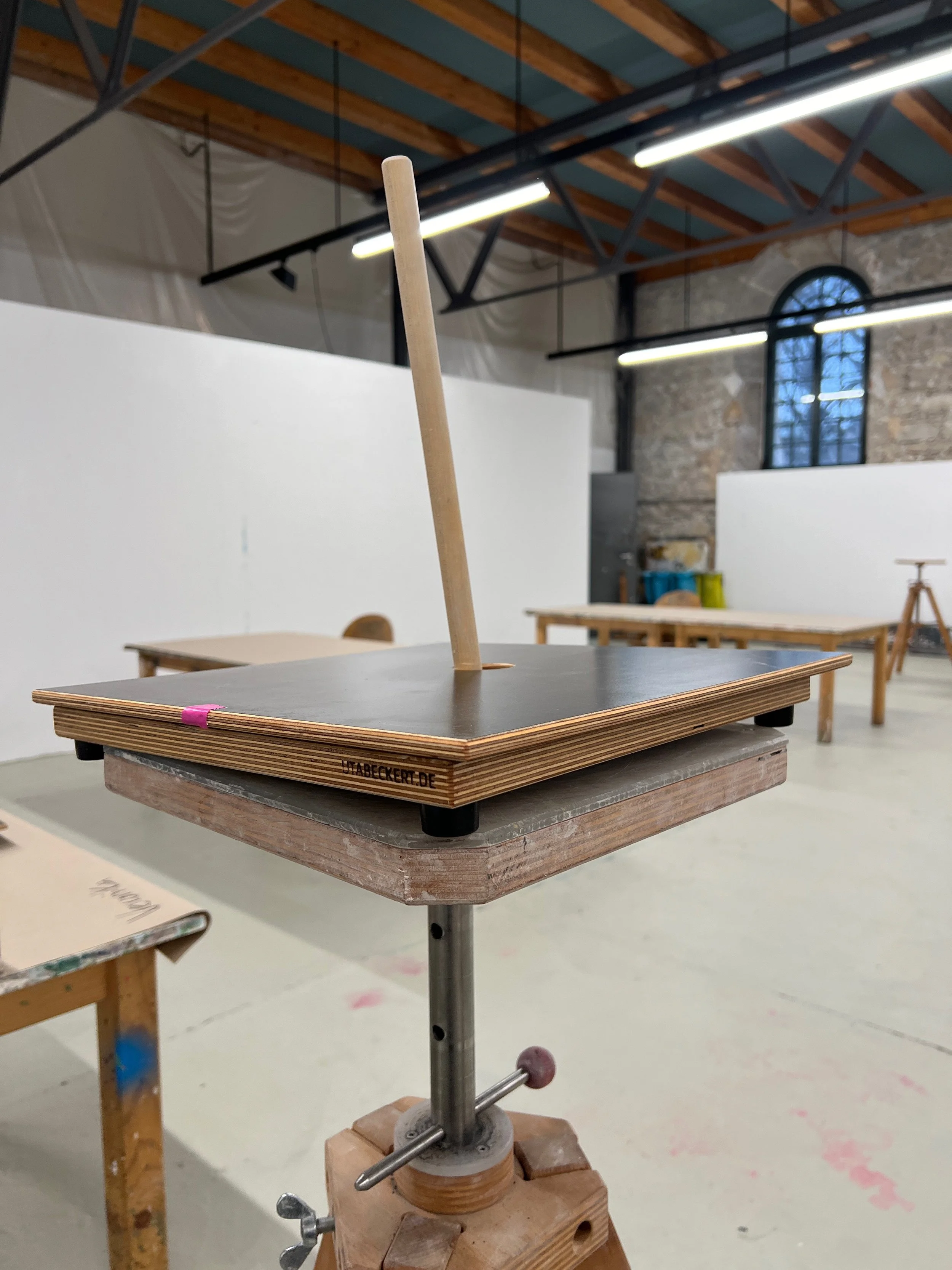 Ein Tisch mit einer seitlichen Klammer, die eine Holzplatte am Tisch festklemmt, in einem Raum mit hohen Decken, Holzbalken, großen Fenstern und weißen Wänden.