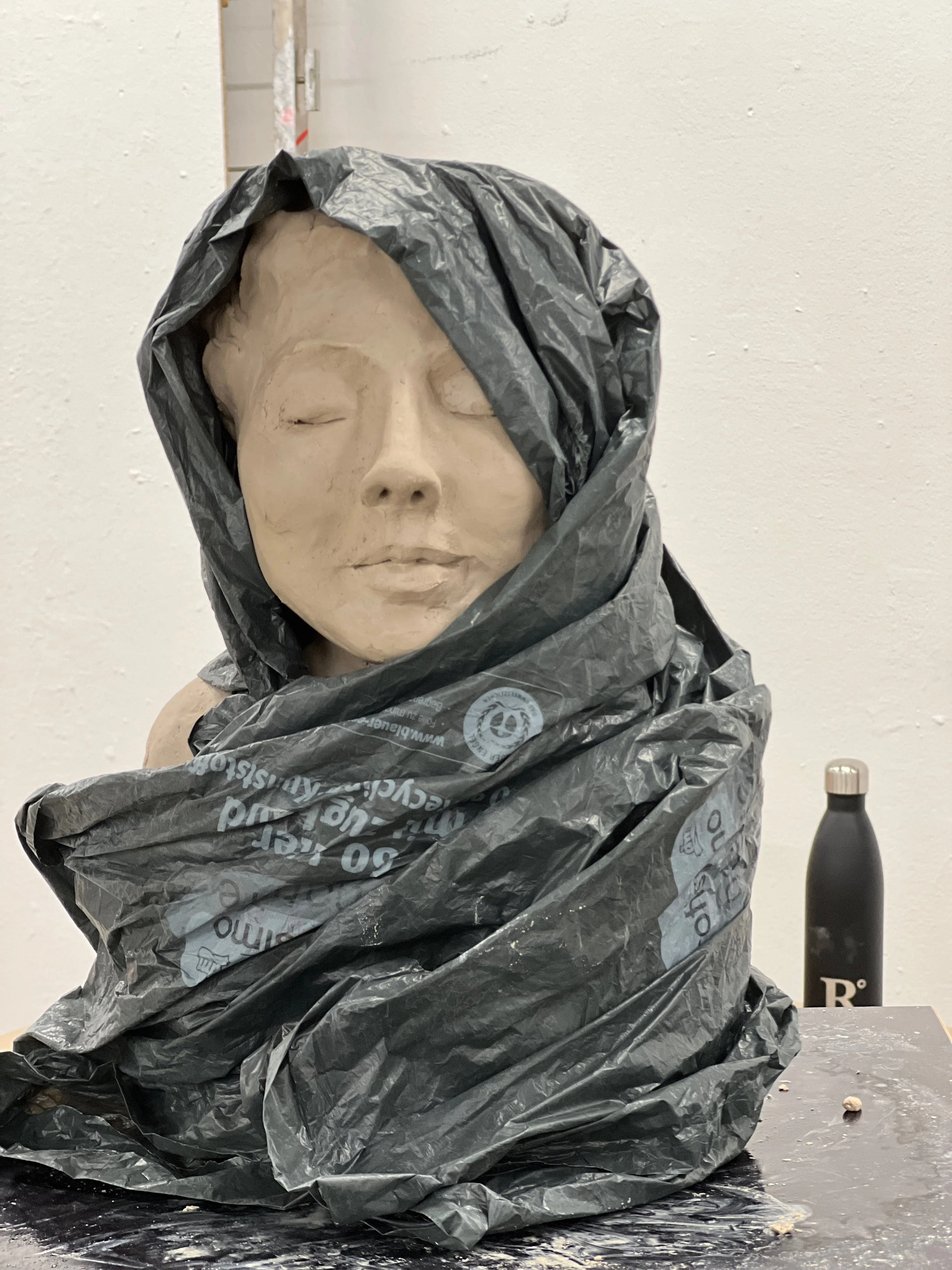 Eine Skulptur eines schlafenden Gesichtes, eingehüllt in eine schwarze Plastiktüte, auf einem Tisch, daneben eine schwarze Wasserflasche.