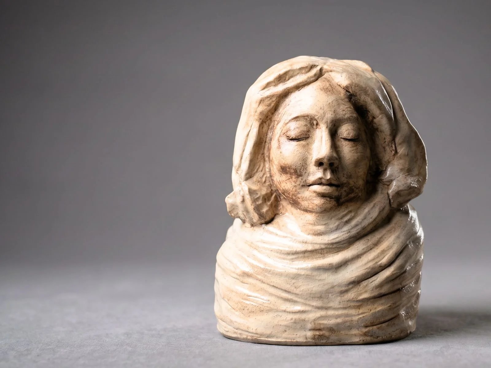 Skulptur eines schlafenden Mädchens mit geschlossenen Augen, umhüllt von einem Stoff, auf grauem Hintergrund.