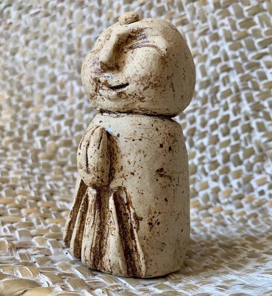 Kleine Skulptur eines lachenden Mannes in einem langen Rock, aus hellem Ton. Im Hintergrund eine strukturierte beige Oberfläche.