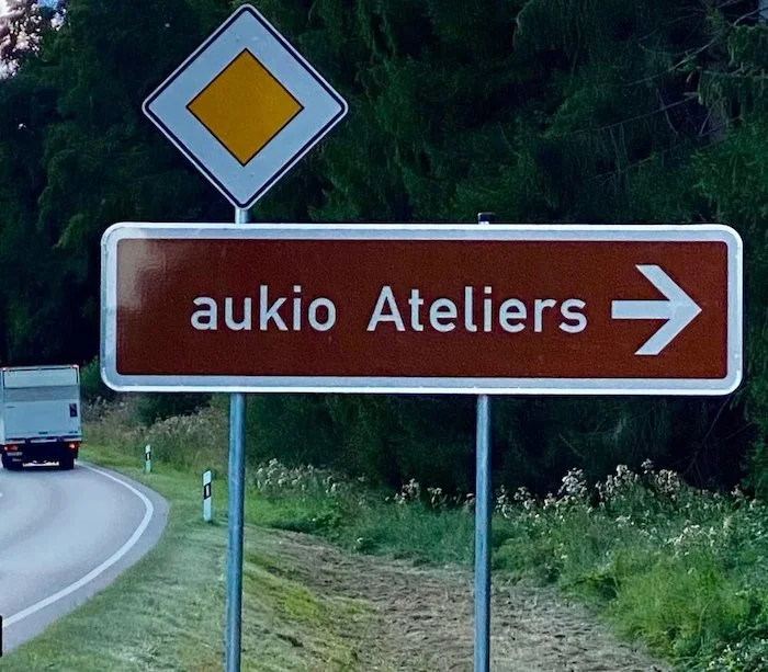 Straßenschild weist nach rechts zu einem Atelier namens "aukio Atelier" auf einer Landstraße, im Hintergrund Bäume und ein Fahrzeug.