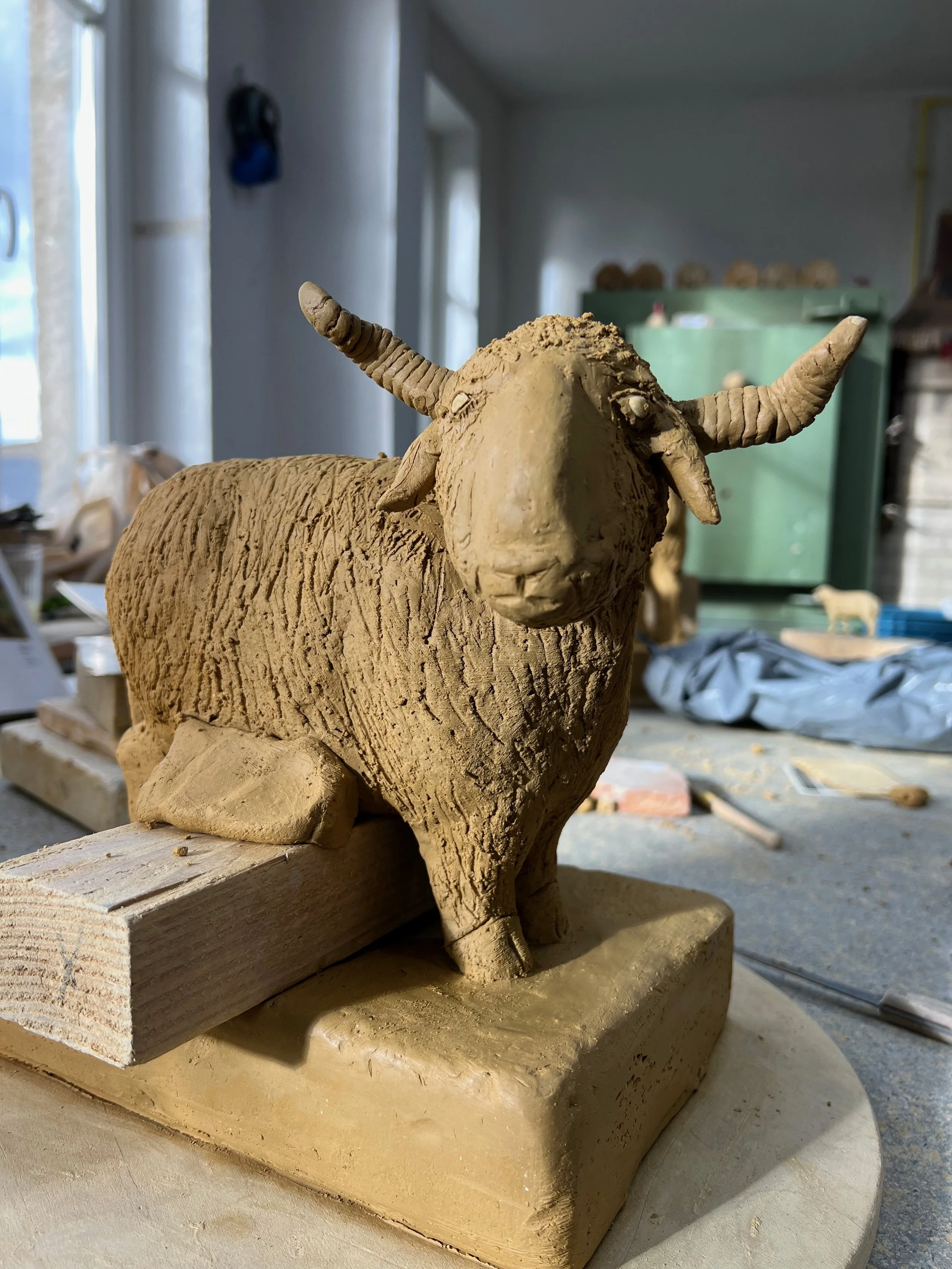 Eine Tierfigur aus Ton oder Ton ähnlichem Material, die ein Büffel oder Wasserbüffel darstellt. Die Figur ist detailliert ausgearbeitet, zeigt den Kopf mit gewellten Hörnern und Textur des Fells, befindet sich auf einem Holzsockel in einem Atelier oder Werkstatt.