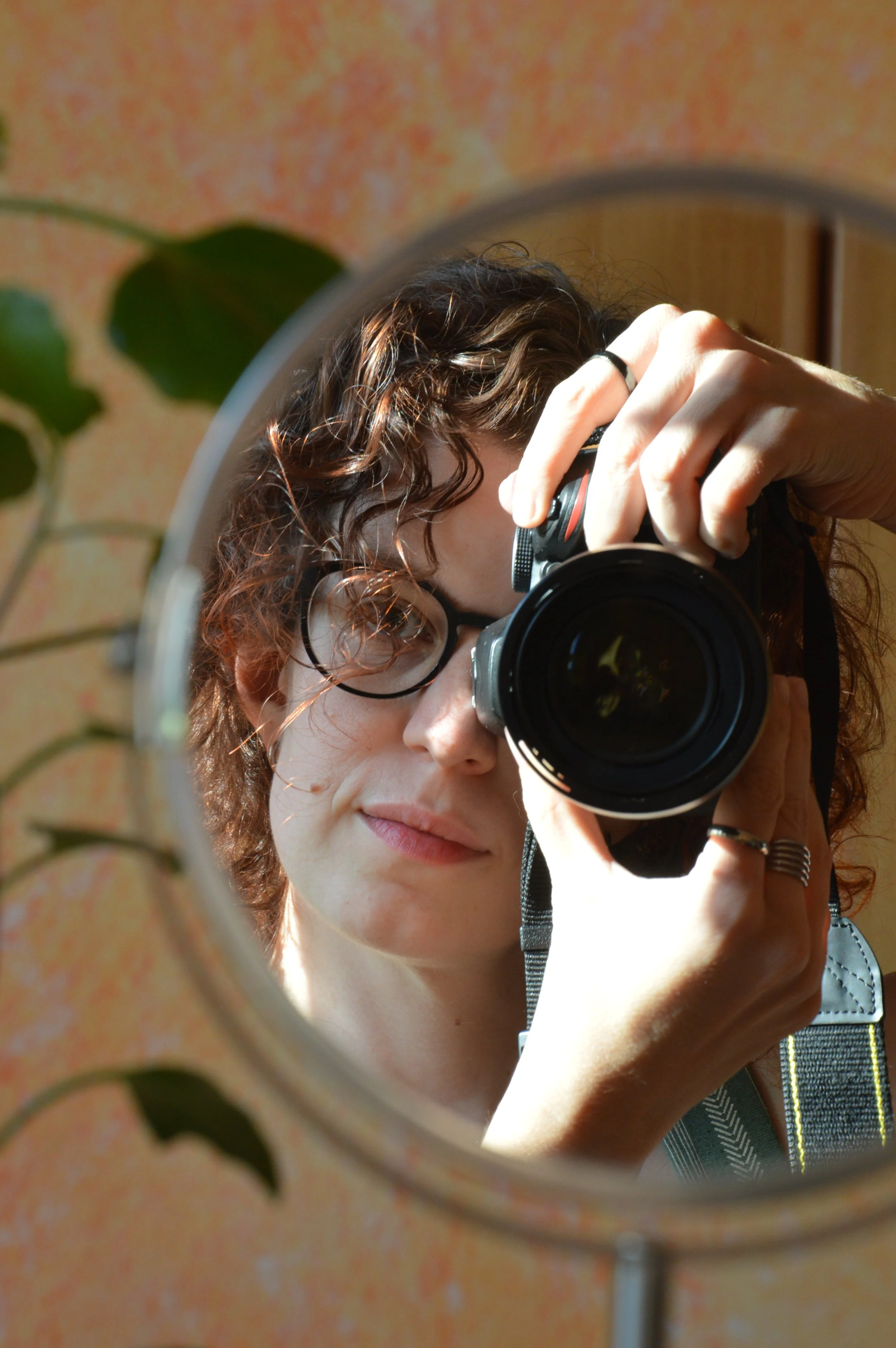 Riflesso di una donna con capelli corti e ondulati che prende una fotografia con una fotocamera reflex, visibile attraverso uno specchio circolare.