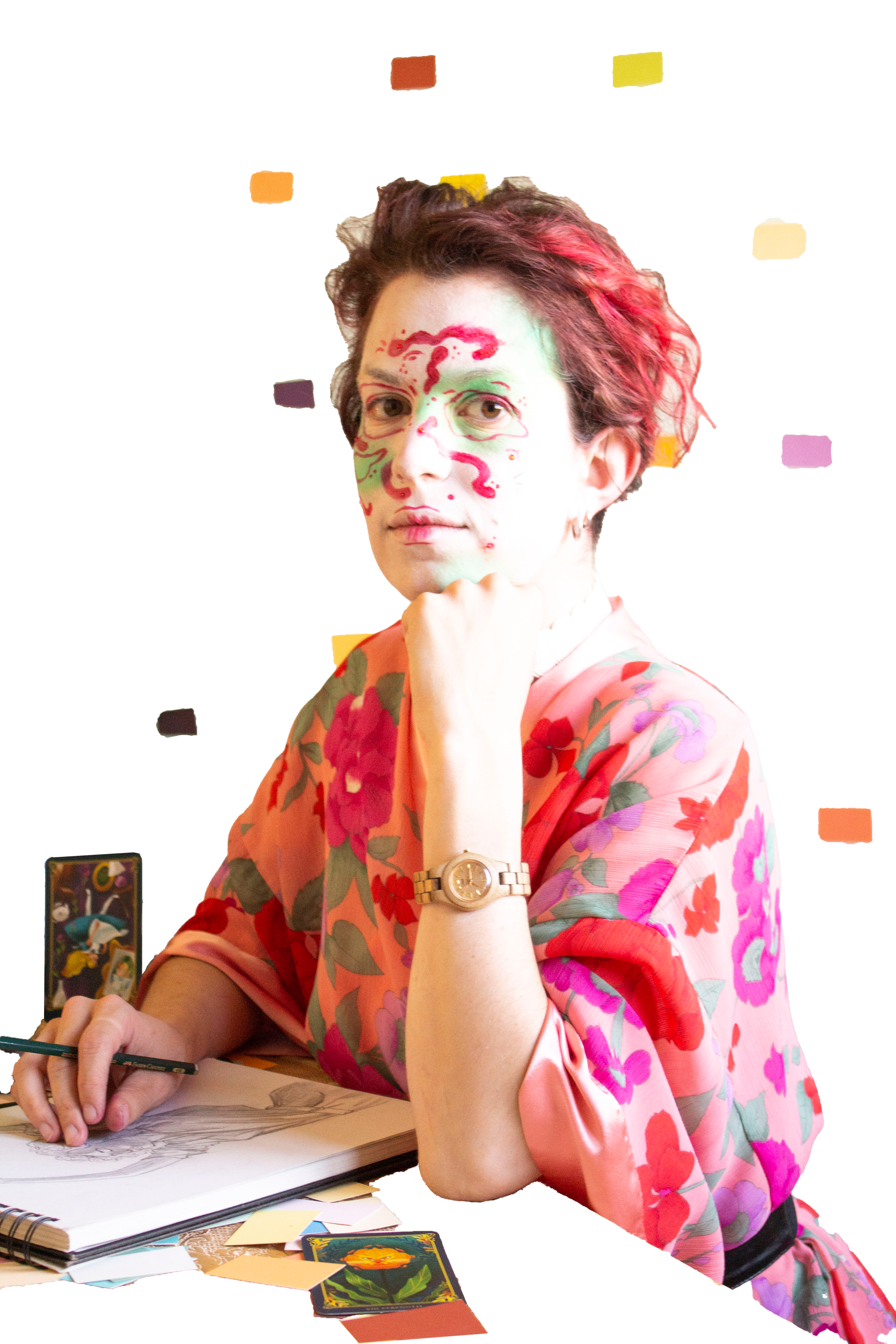 L'illustratrice con capelli ricci e trucco artistico sul viso, indossa un kimono rosa a fiori, seduta mentre disegna su un quaderno, guarda in camera ed circondata da carte colorate e tarocchi.