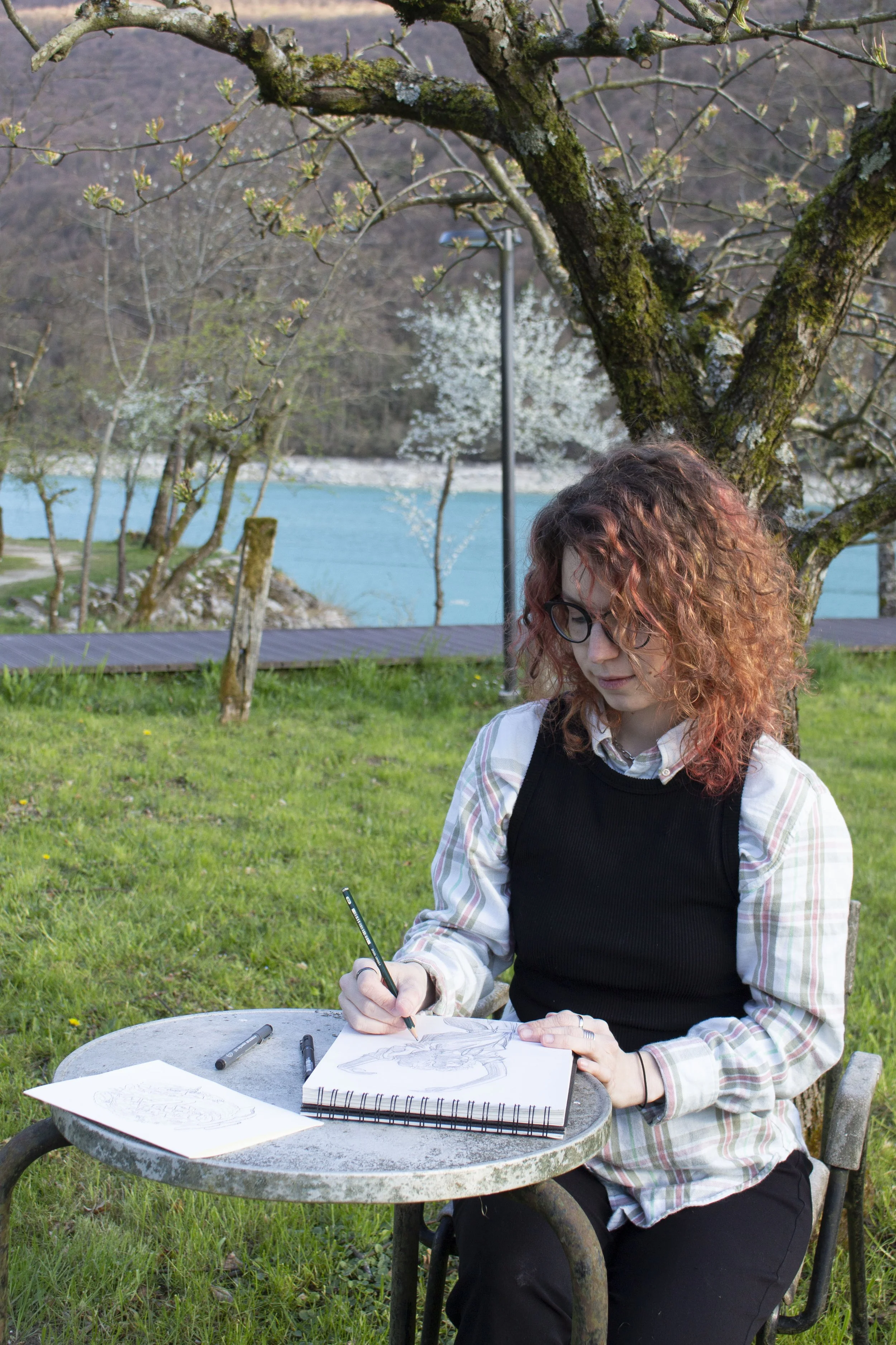 Una donna con capelli ricci rossi, indossa occhiali, una maglia a quadri e un top nero e sta disegnando in uno sketchbook all'aperto su un tavolo di pietra rotondo. Sul tavolo ci sono alcuni pennarelli e un foglio con un disegno. Sullo sfondo si vede un albero con rami spogli e un paesaggio di montagna, un lago e alberi in fiore.