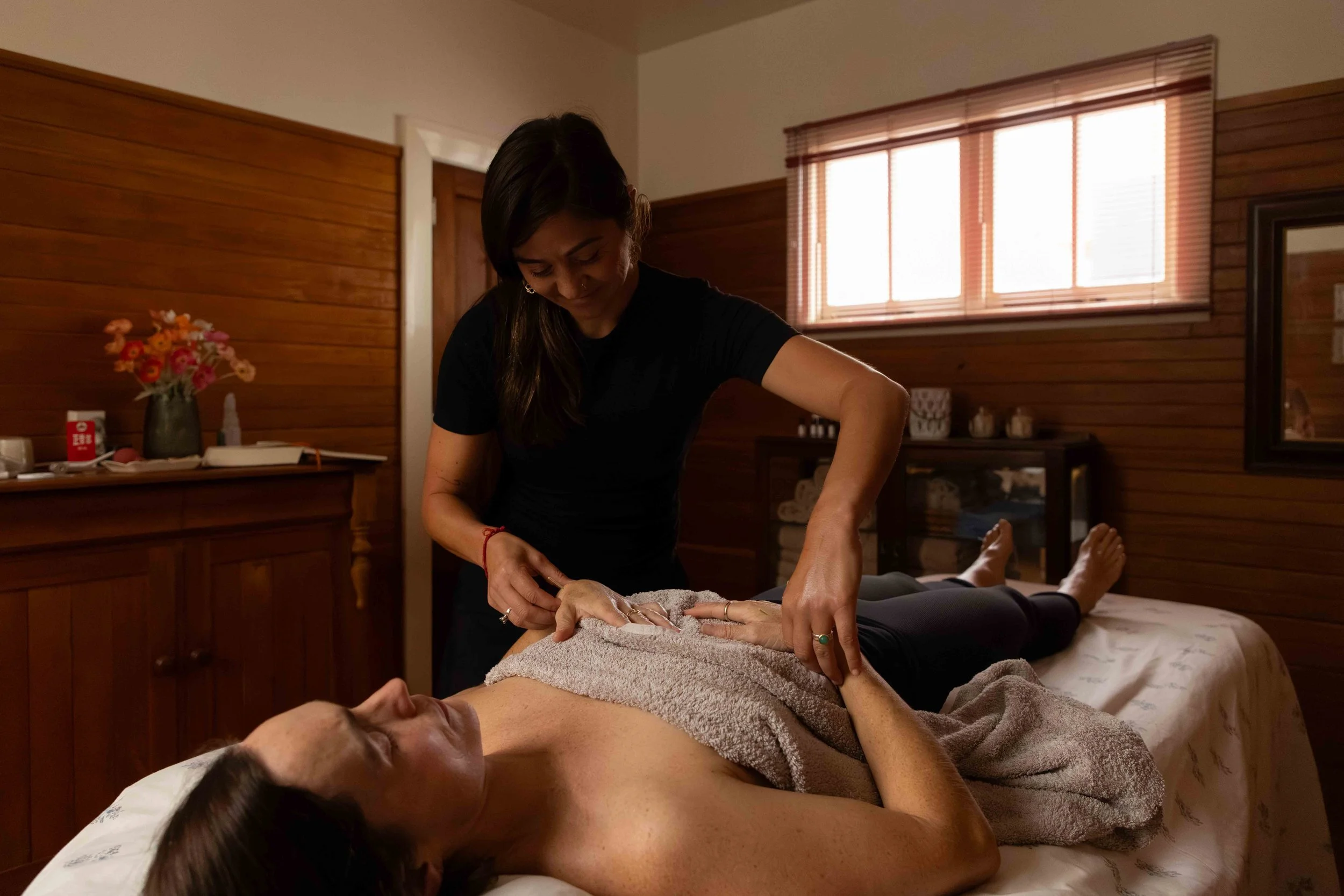 A woman giving a acupuncture treatment to a person lying on a massage table in a cozy, wooden-paneled room with a window.