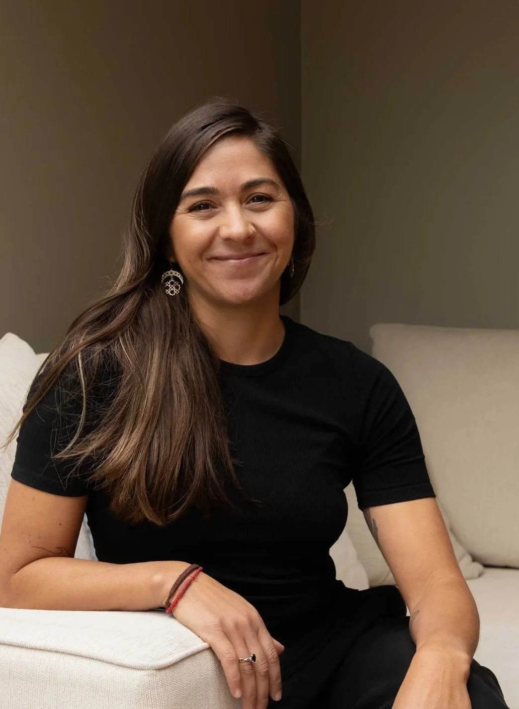 A woman with long brown hair, wearing a black shirt, earrings, and a ring, sitting on a white couch with a smile.