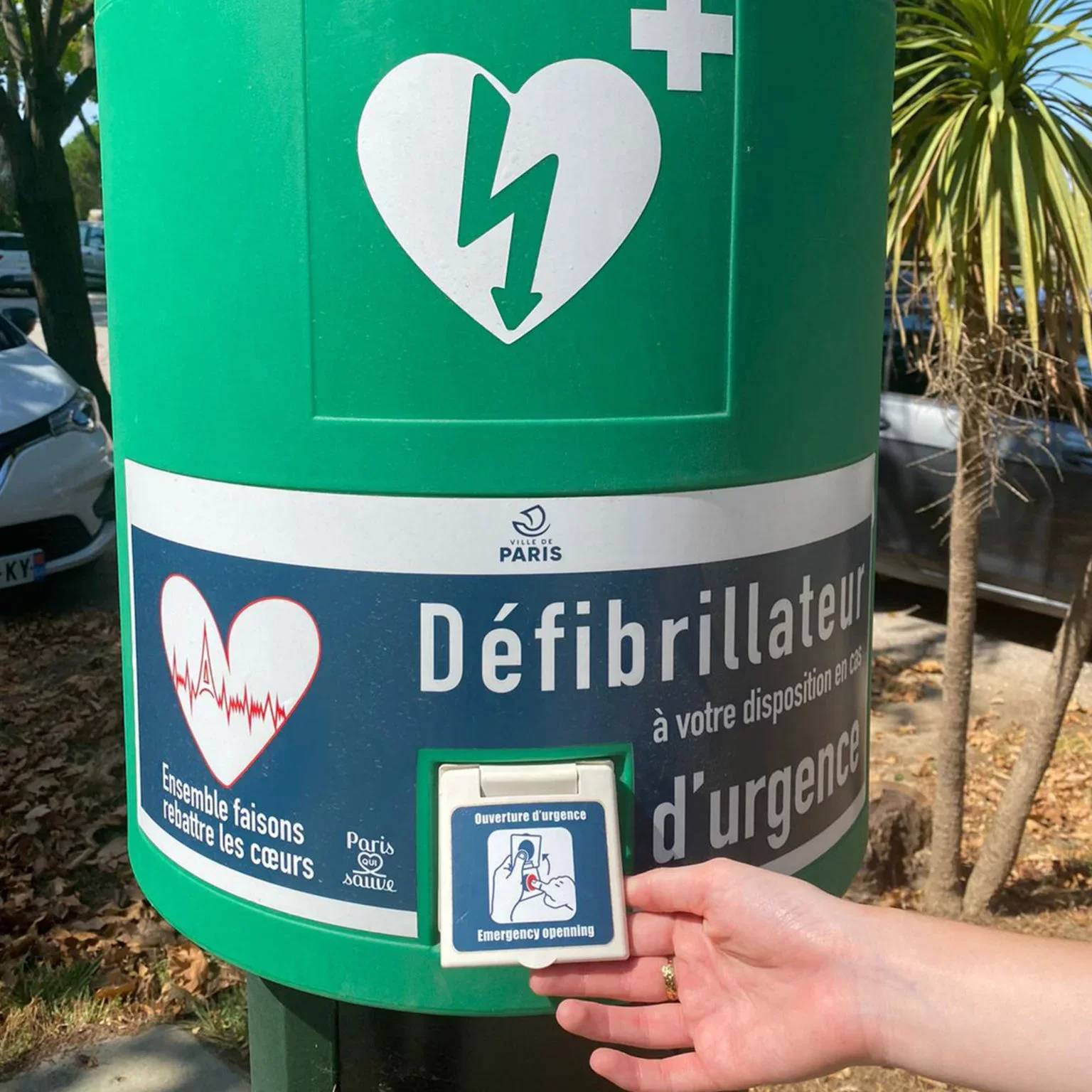 Une personne ouvre une boîte d'urgence pour réanimer un cœur, située à Paris, avec un symbole de cœur avec un éclair et un défibrillateur. La boîte est verte avec des instructions en français et en anglais.