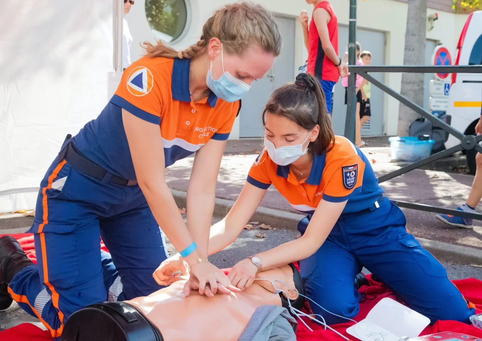 Deux femmes en uniforme de protection CIVI, en train de pratiquer une réanimation sur un mannequin de formation, toutes deux portant un masque médical. Elles semblent être dans un contexte de formation ou d'intervention d'urgence.