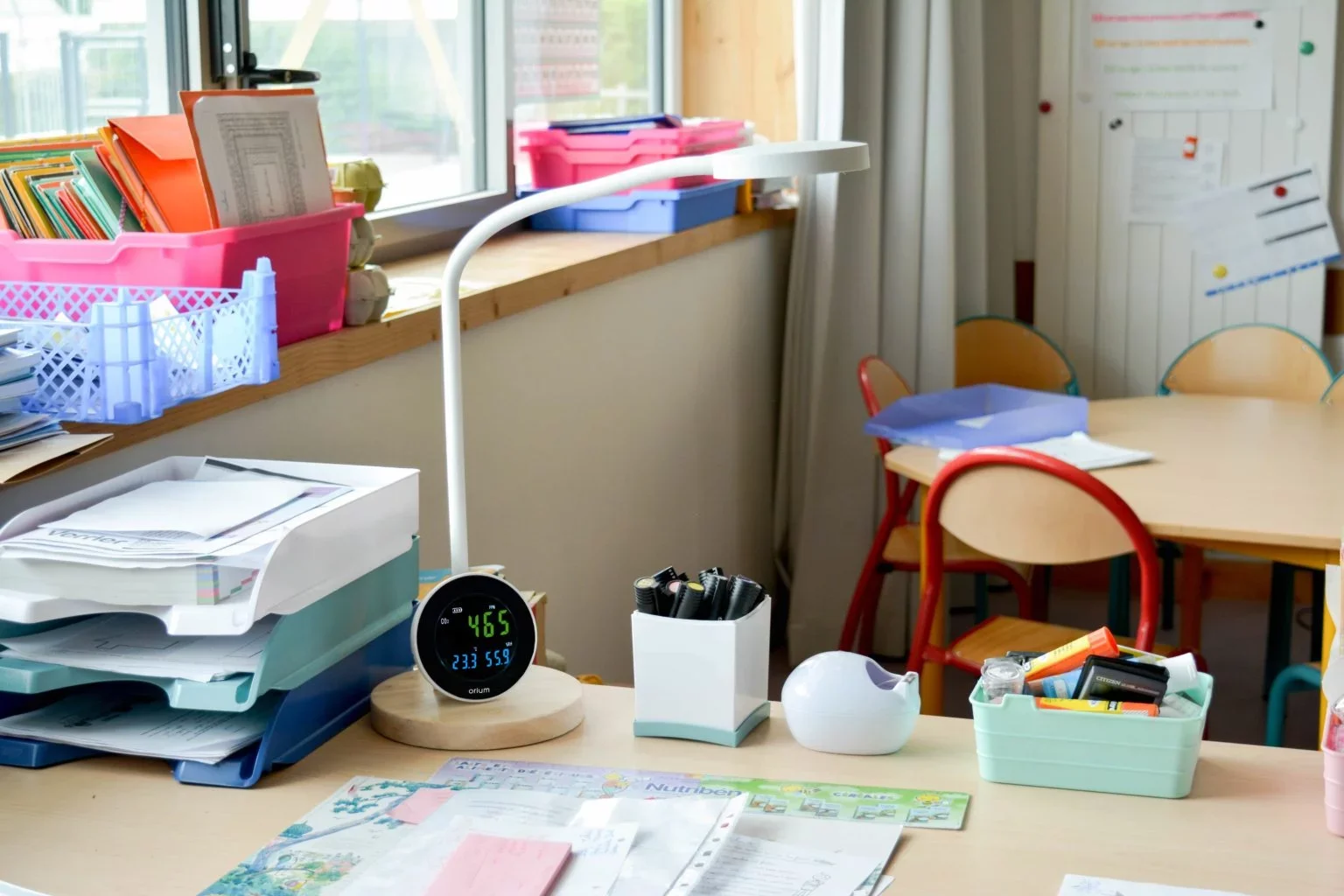 Un bureau de classe avec des piles de papiers, des classeurs, des stylos, un thermomètre numérique dans un support en bois, et des chaises colorées.