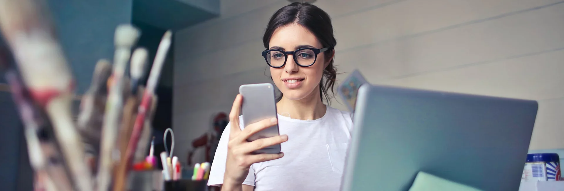 Une jeune femme portant des lunettes regarde son téléphone portable, avec un ordinateur portable devant elle et un pot de crayons à la main.