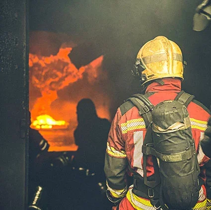 Un pompier regarde un incendie à l'extérieur, avec des flammes et de la fumée orange dans le feu.