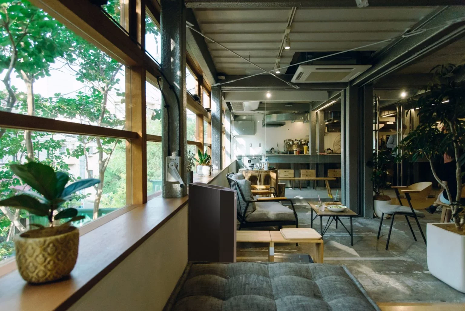 Intérieur d'un café avec des fauteuils, tables, plantes en pot, grandes fenêtres laissant entrer la lumière naturelle, et une cuisine en arrière-plan.