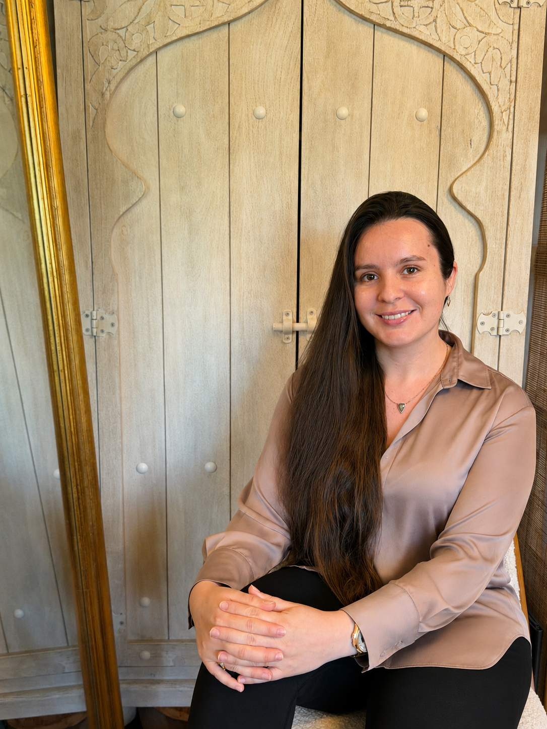 A woman with long dark hair sitting in front of a wooden, carved wardrobe, wearing a beige satin blouse and black pants, smiling at the camera.
