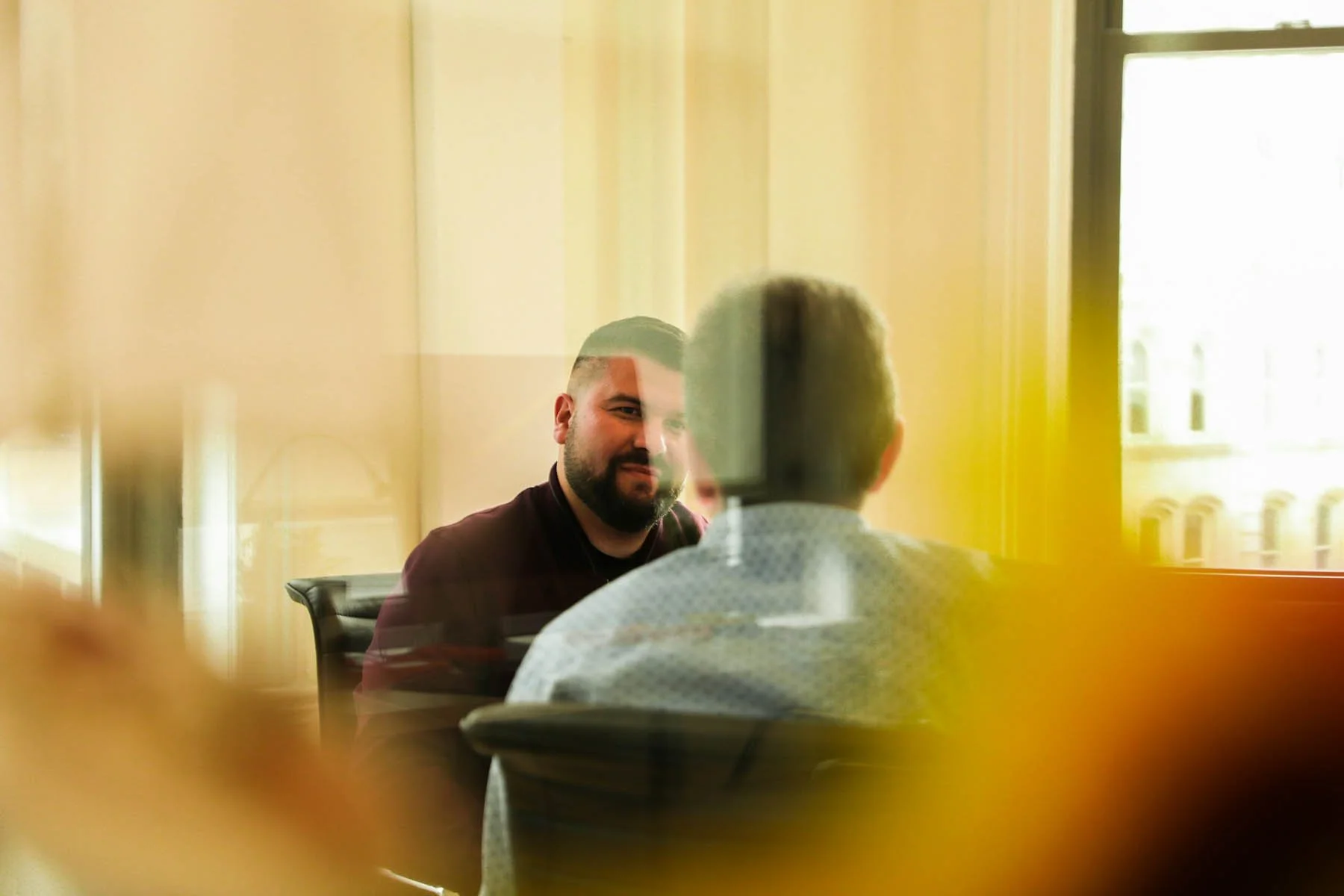 Two men sitting and talking in a room with yellow walls and large windows, viewed through a window with reflections and an out-of-focus foreground.