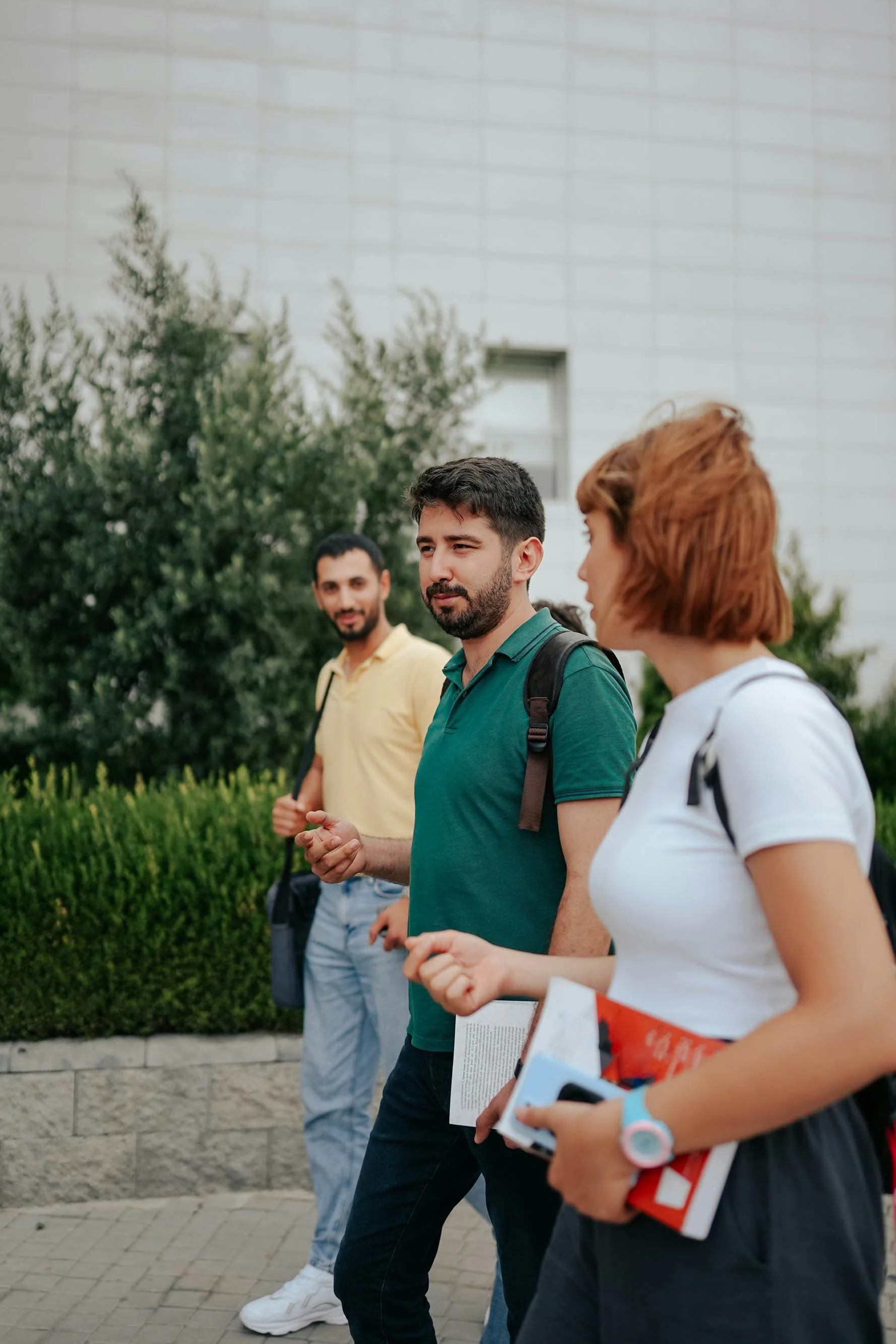 Three young adults smile and walk outdoors with greenery and a white building wall in the background. They are casually dressed and appear to be engaged in conversation.
